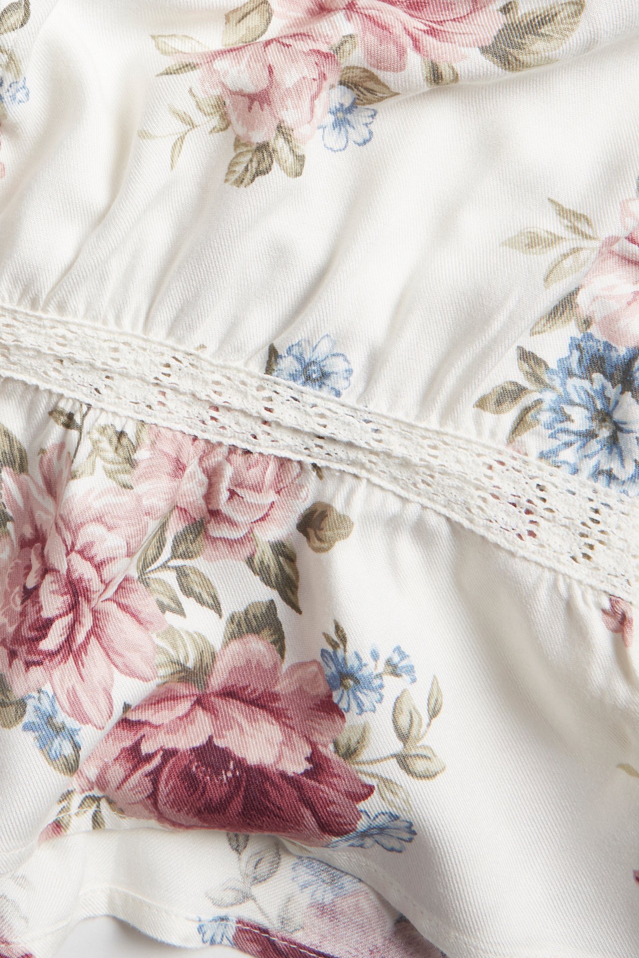 Nahaufnahme einer cremefarbenen floralen Viskosebluse mit rosa Rosen, blauen Blüten und zarter Spitze.