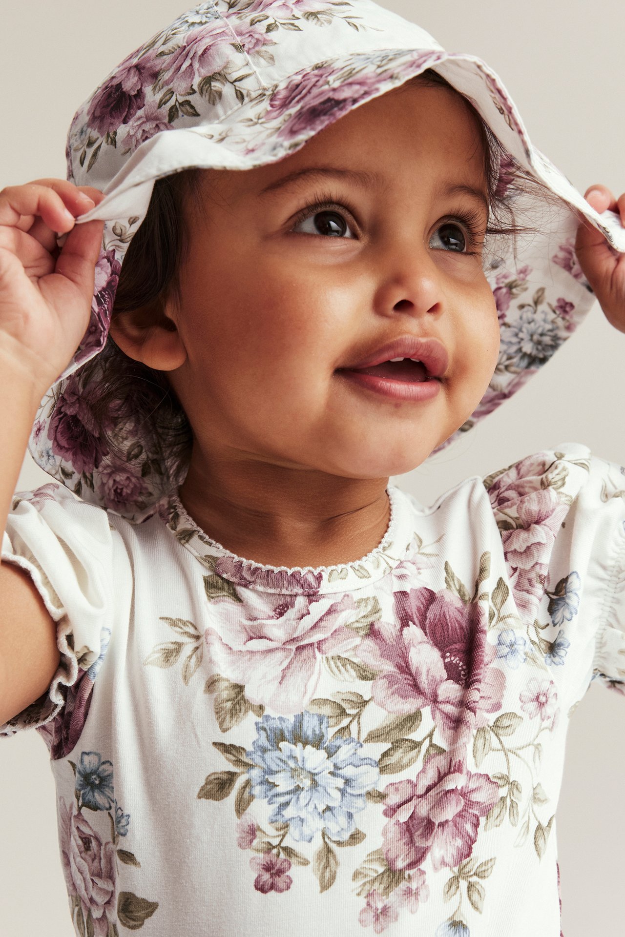 Close-up of a baby wearing an off-white floral sun hat with a wide brim and a matching floral top.