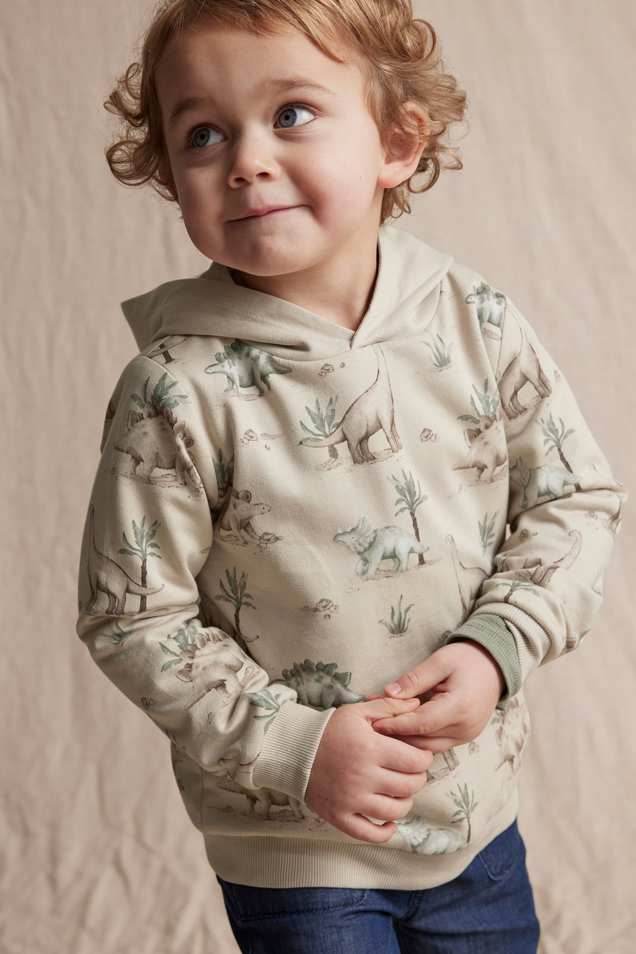 Front view of a kid wearing a beige cotton hoodie with a dinosaur pattern and blue jeans.