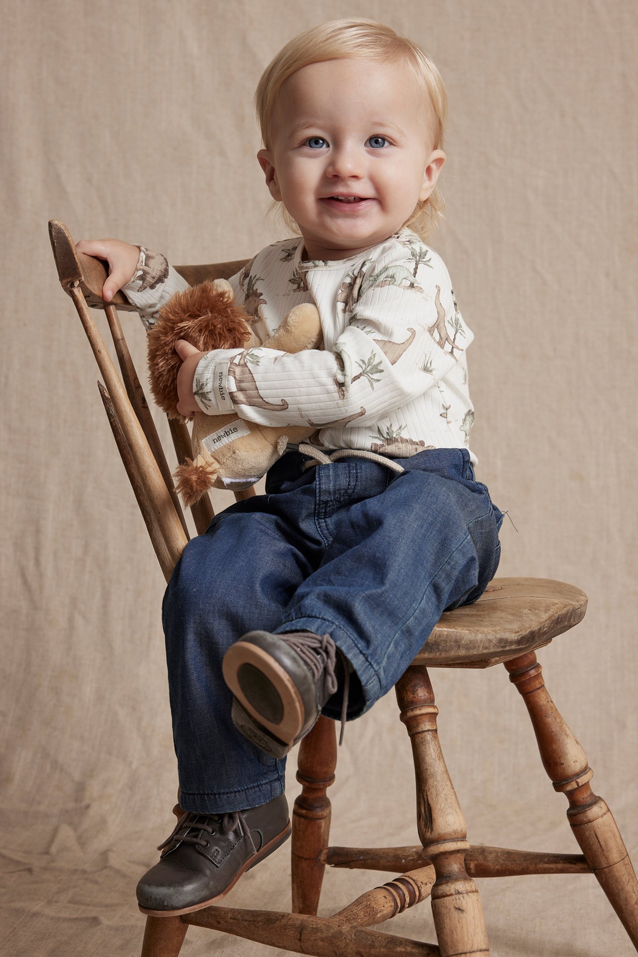Front view of a baby in an off-white ribbed dinosaur bodysuit, blue trousers, and brown shoes, holding a toy.