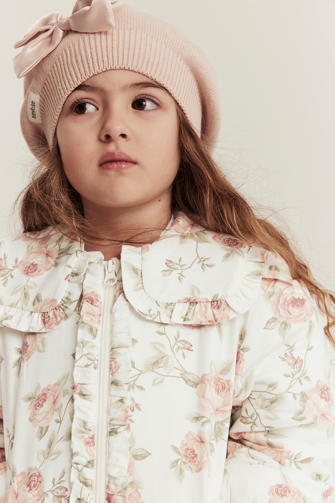 Close-up of a kid wearing an off-white floral bomber jacket with ruffles and a light pink beret with a bow.