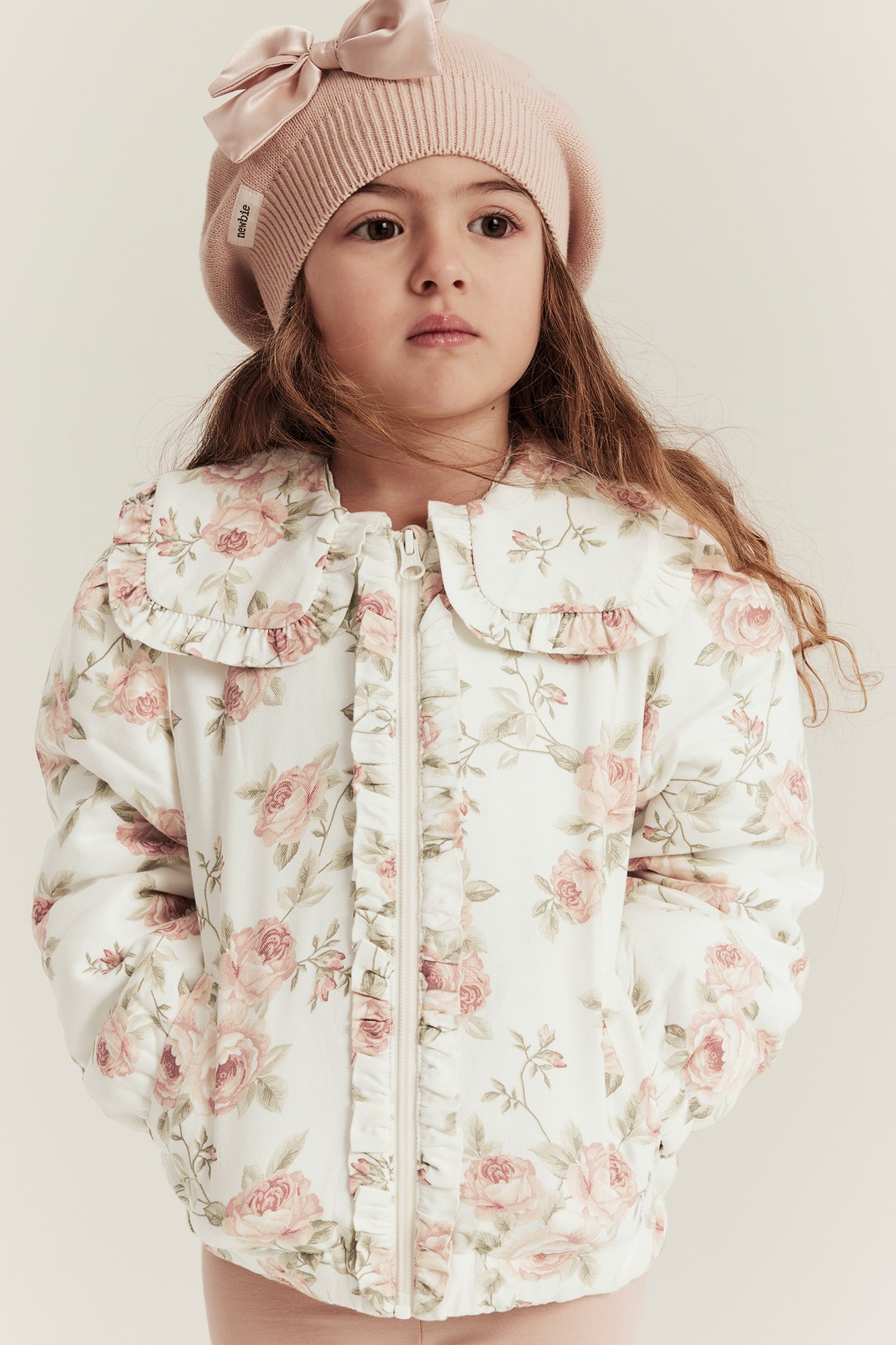 Front view: kid girl in an off-white floral bomber jacket with ruffles and a pink knitted beret with a bow.