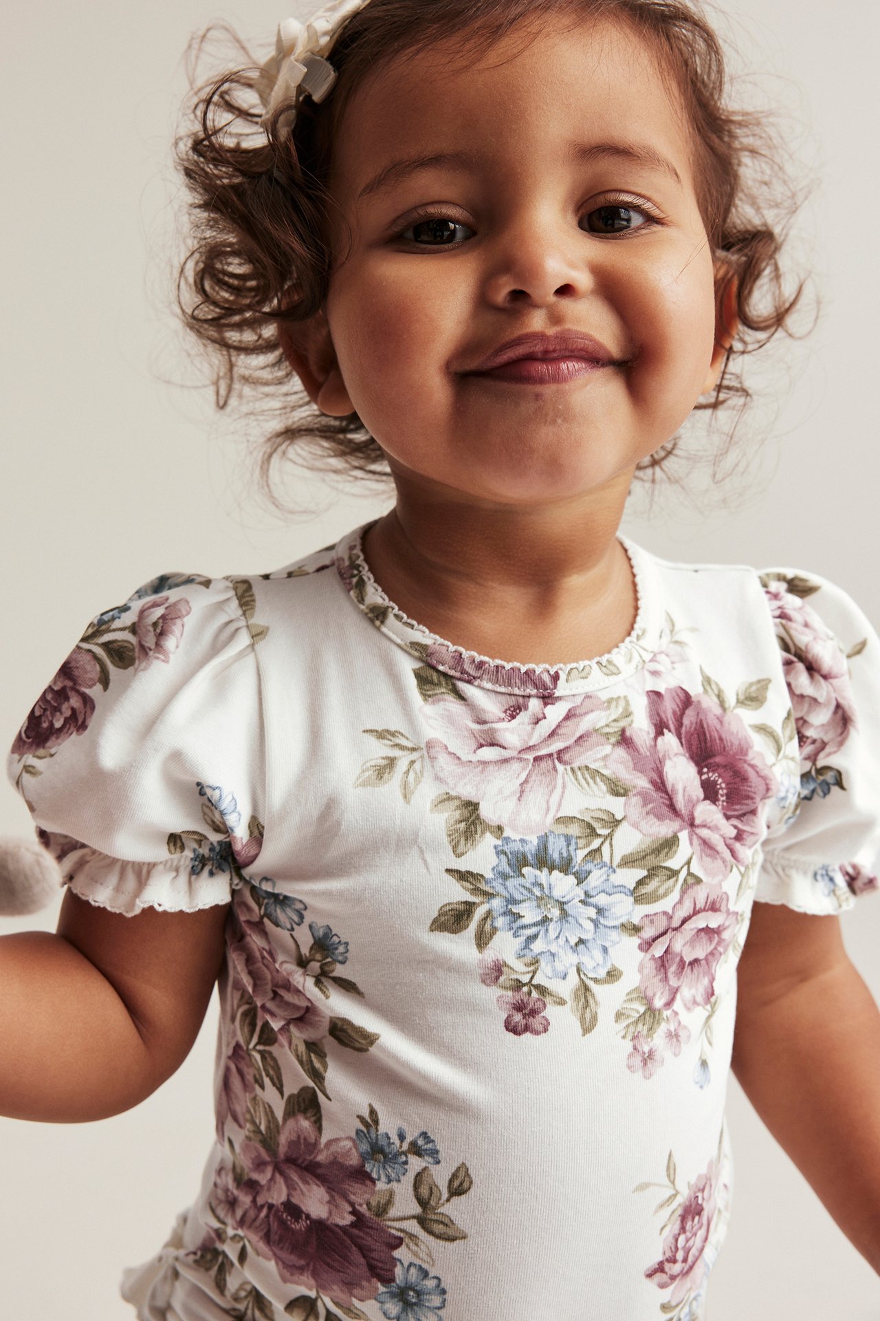 Close-up front view of a baby wearing an off-white floral puff-sleeve bodysuit with picot trim and a hair bow.