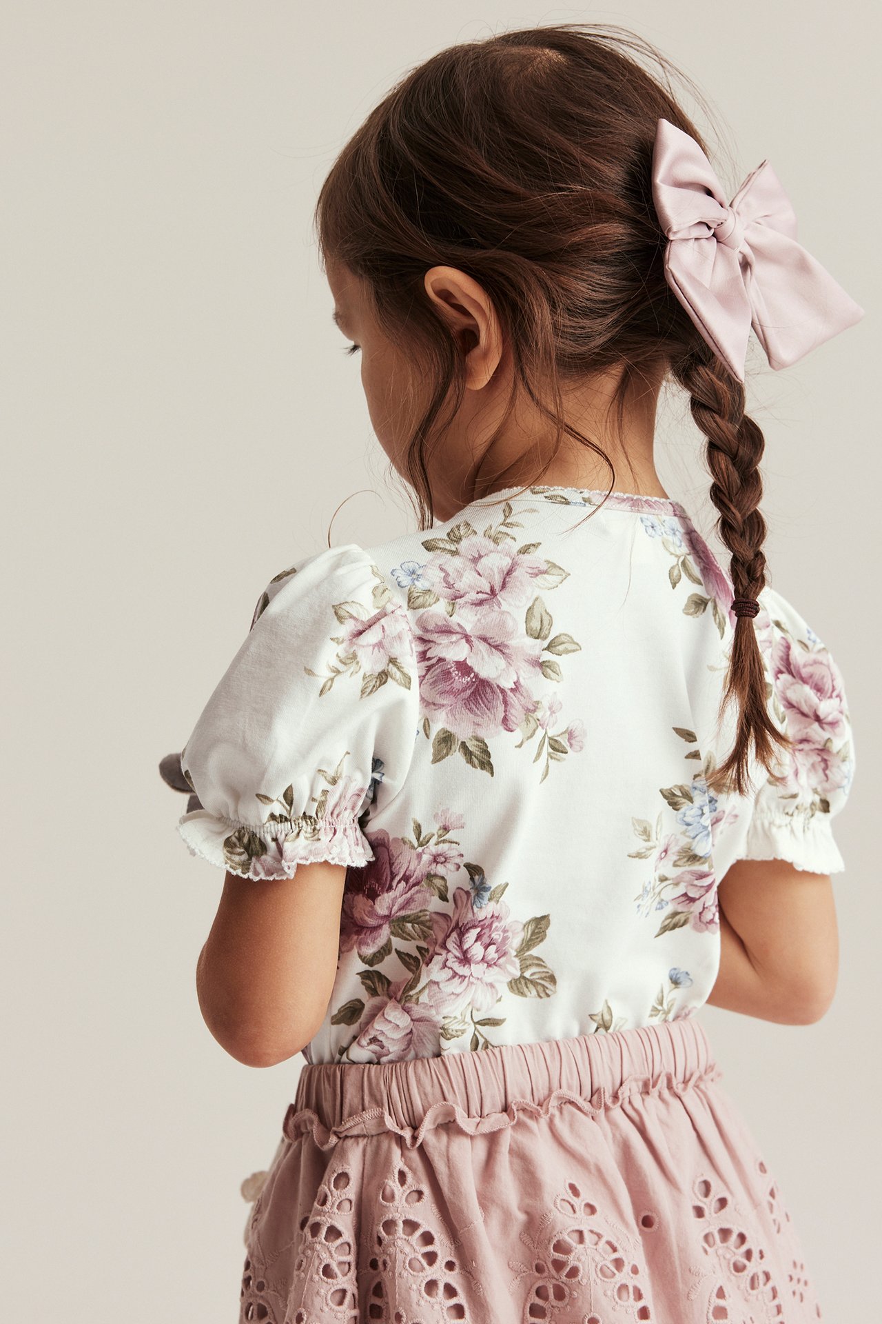 Rückansicht: Kind in cremefarbenem Blümchen-Puffärmel-Oberteil, rosa Lochstickerei-Rock und rosa Haarschleife.