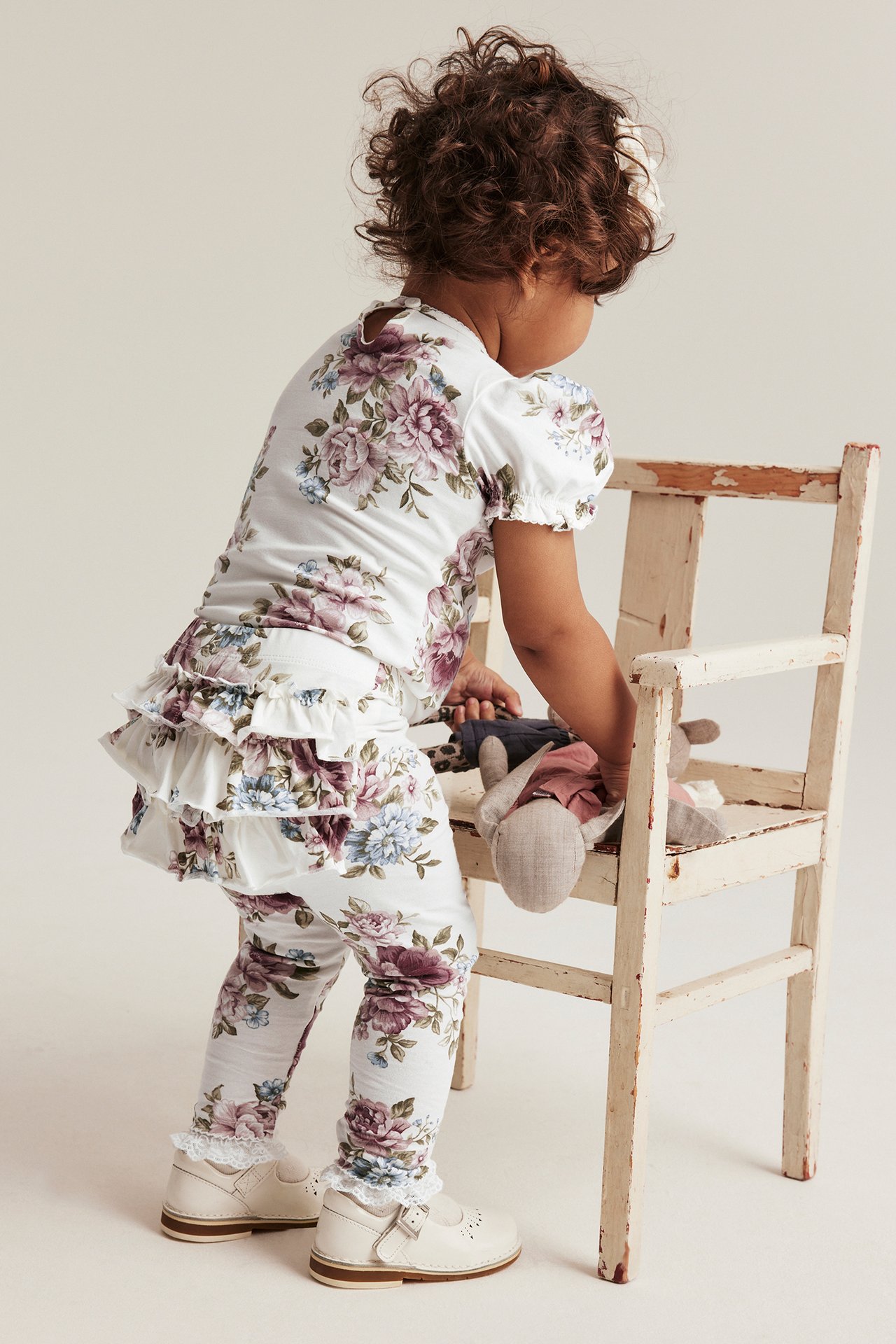 Back view of a baby in off-white floral ruffled leggings, matching top, and off-white shoes, playing with a doll.