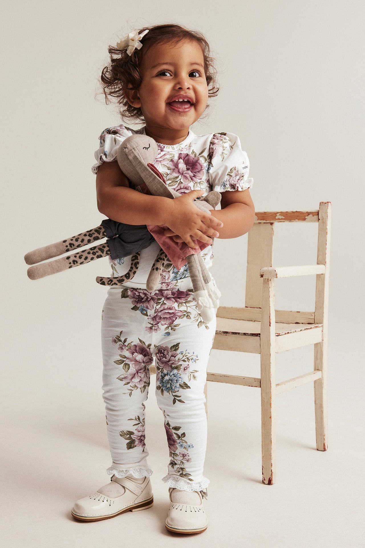 Front view: baby girl in offwhite floral leggings with ruffles, matching top, offwhite shoes, holding a toy.