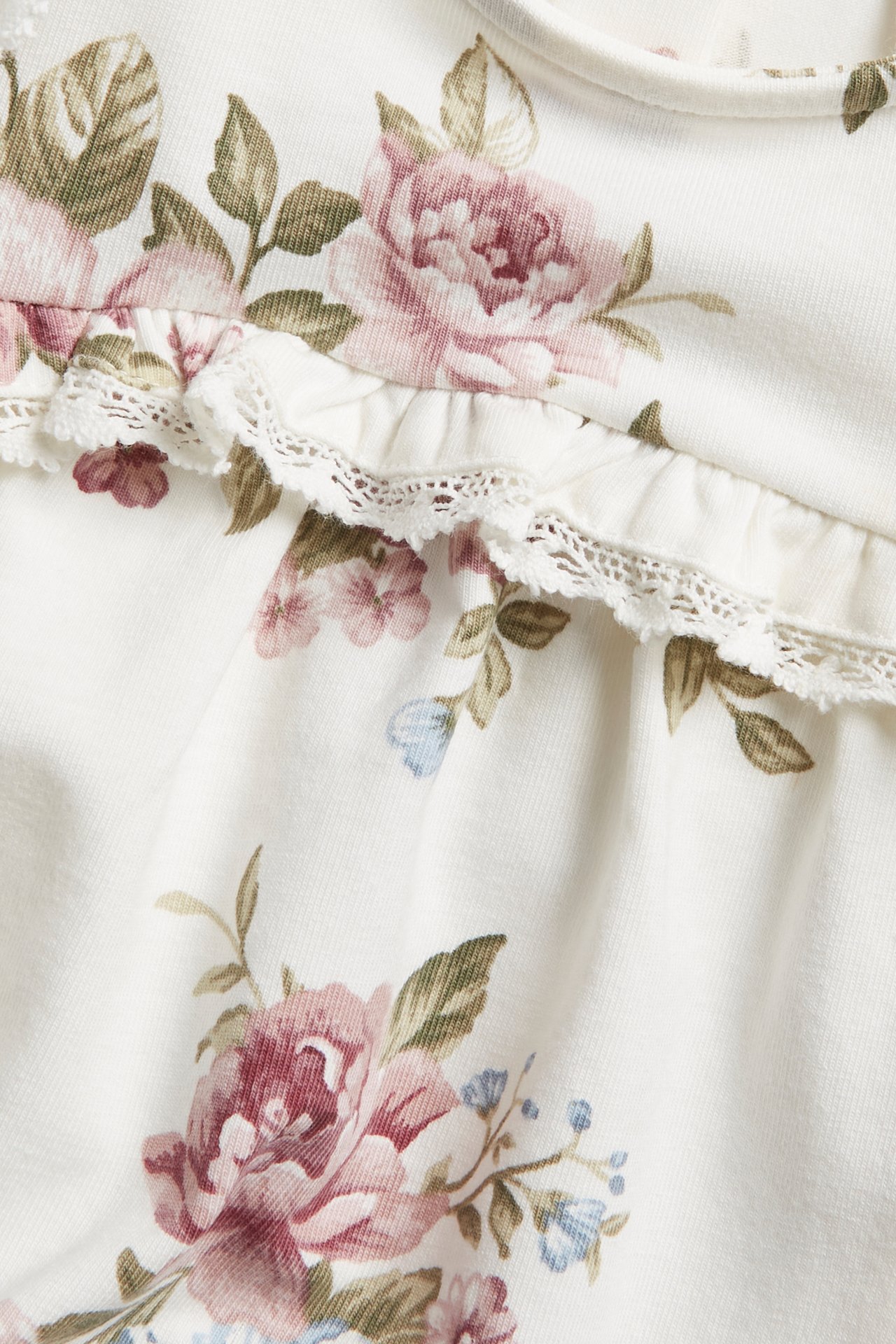 Close-up of an off-white floral jersey dress with pink roses, green leaves, ruffles, and white lace trim.