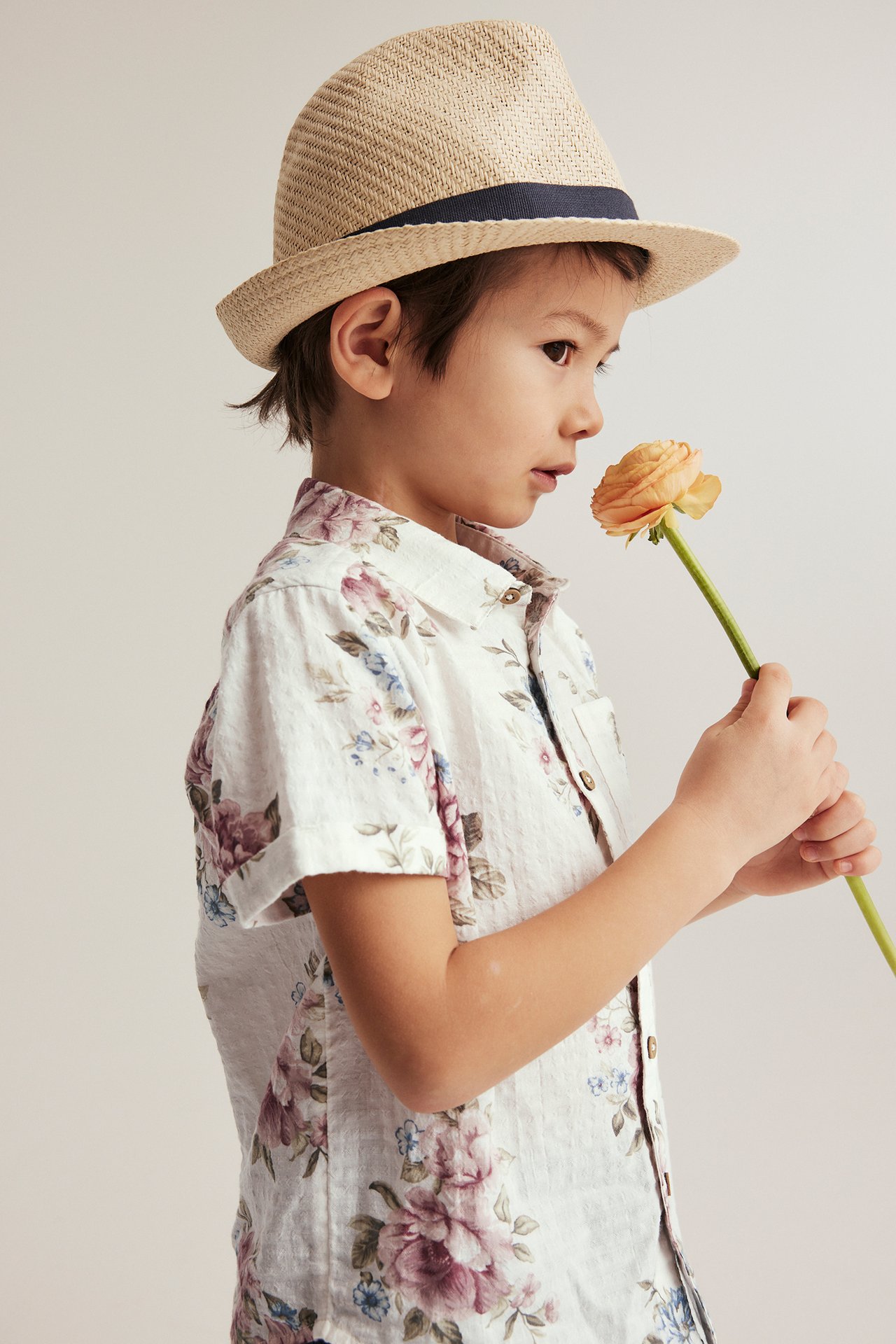 Side view of a kid in an off-white floral short-sleeved cotton shirt, beige straw hat, holding an orange flower.