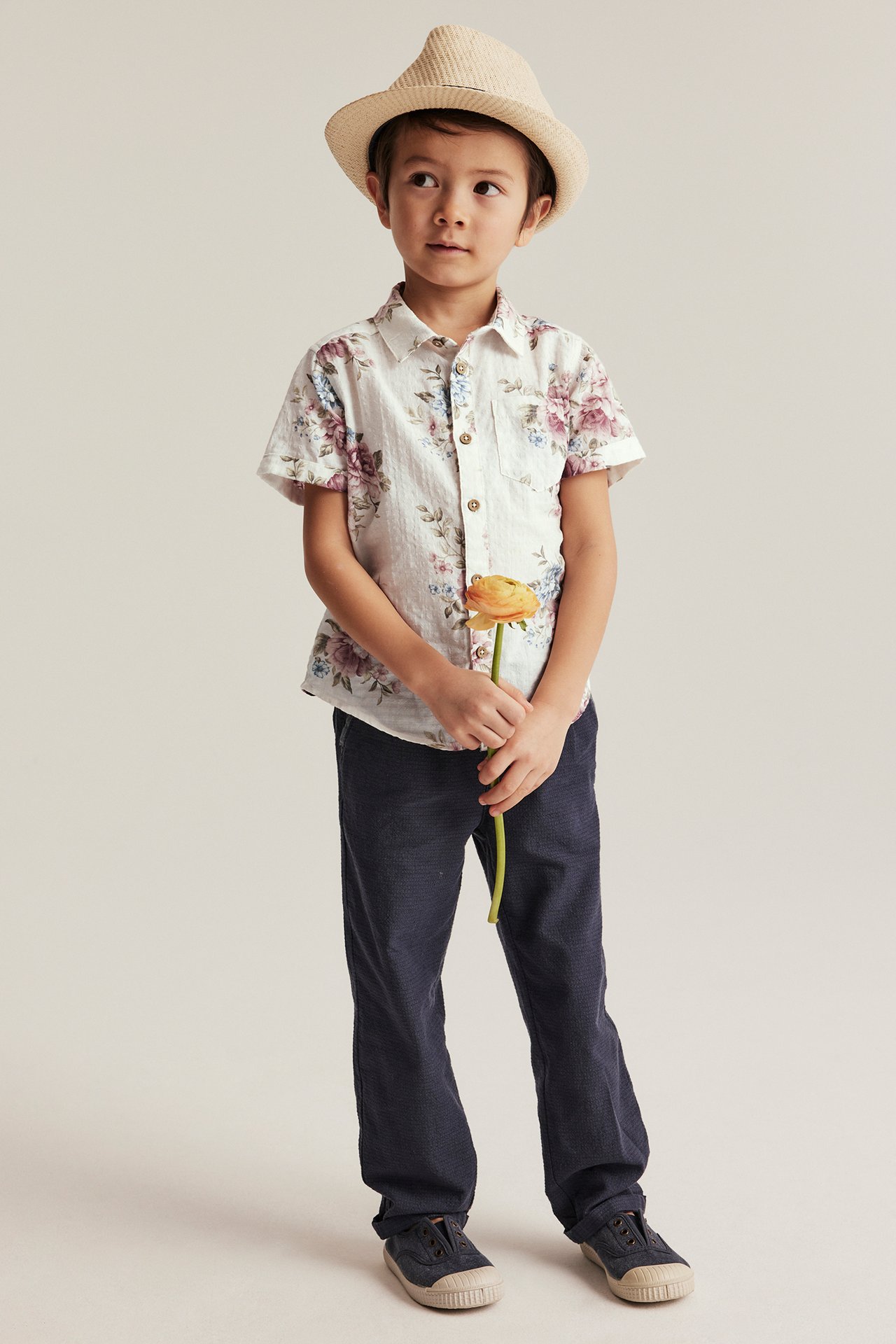 Front view: Kid in off-white floral short-sleeved cotton shirt, dark blue trousers, slip-on shoes, straw hat.