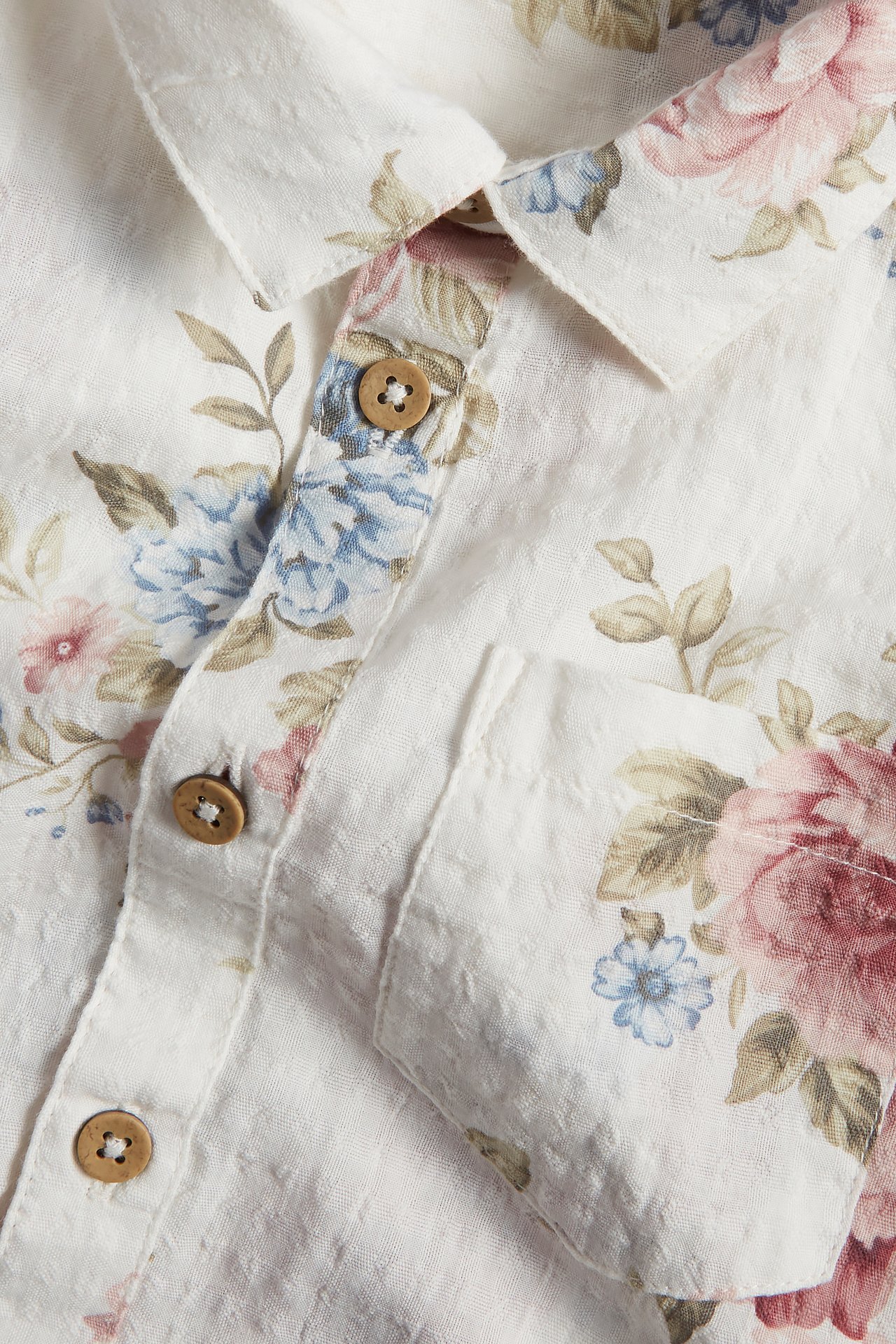 Close-up of an off-white short-sleeved cotton shirt with floral pattern, wooden buttons, front pocket.