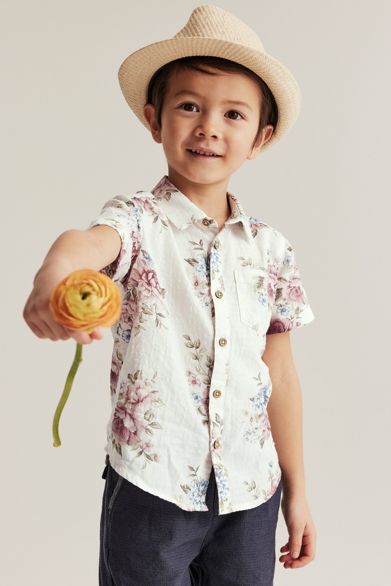 Front view: Kid in off-white floral short-sleeved shirt, dark trousers, straw hat, holding orange flower.