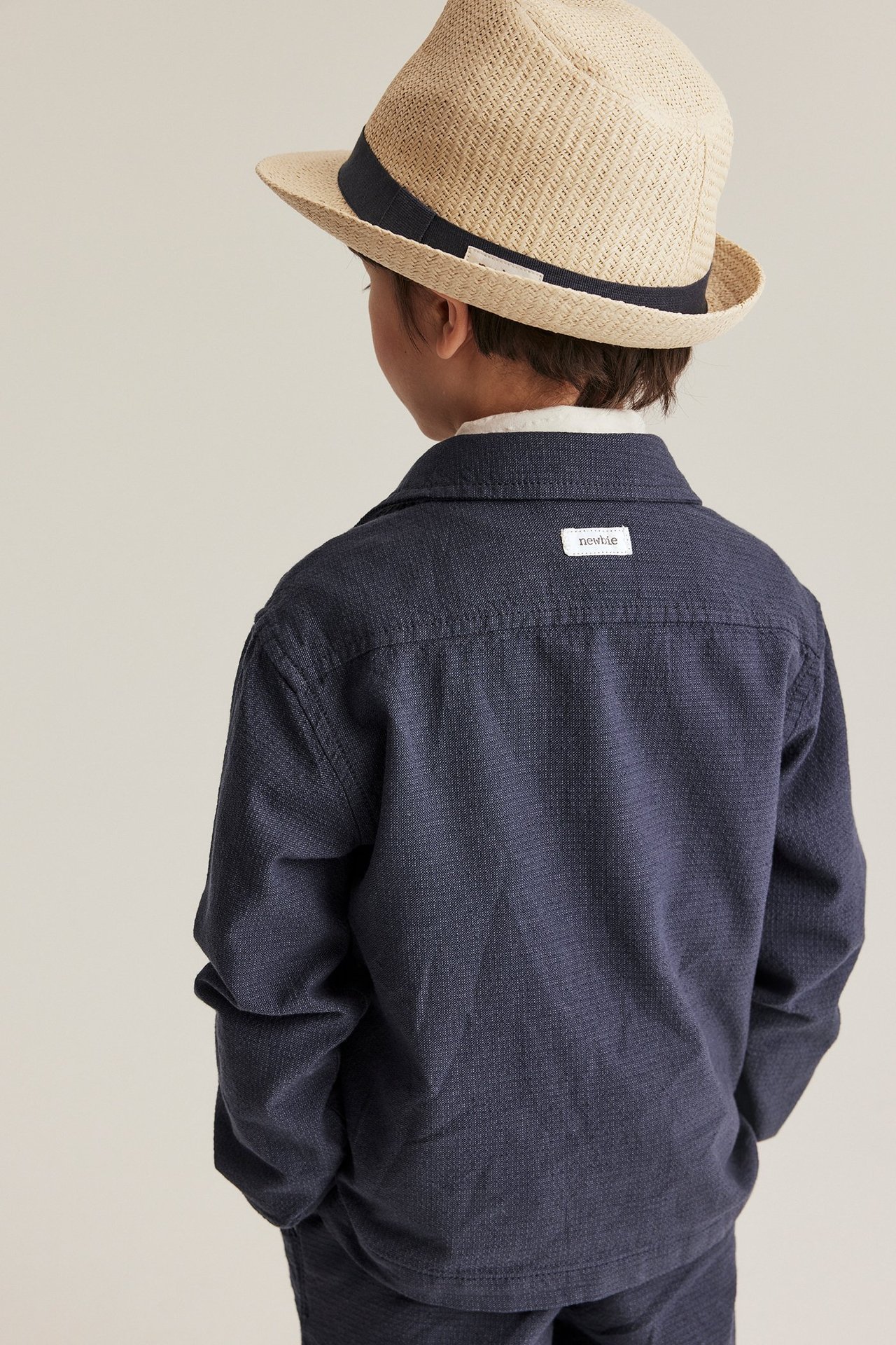 Rückenansicht: Kind in dunkelblauem Chambray-Overshirt und beigem Strohhut mit dunklem Band.