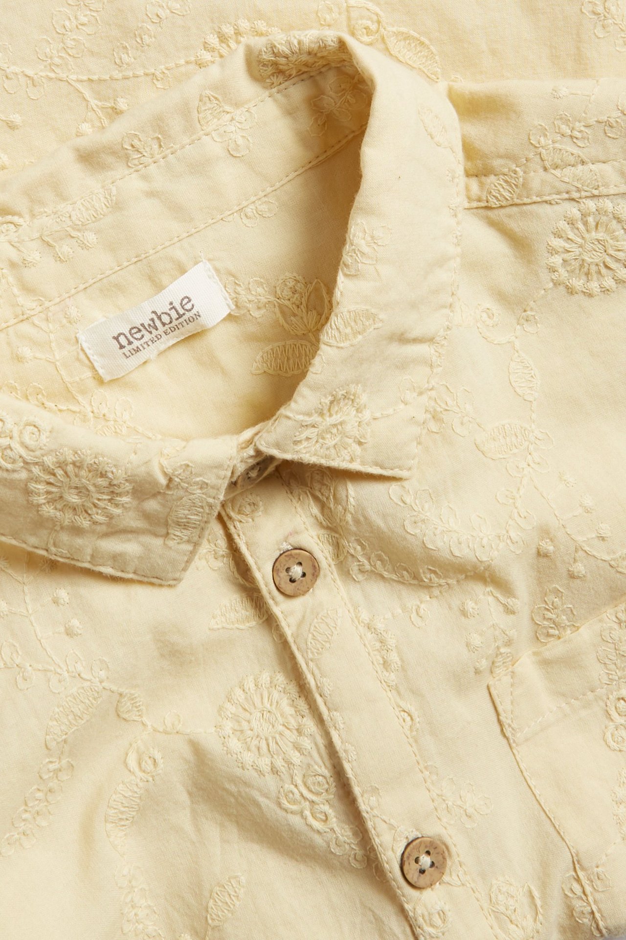 Nahaufnahme: gelbes kurzärmeliges Baumwollhemd mit Blumenstickerei, Kragen und Holzknöpfen.