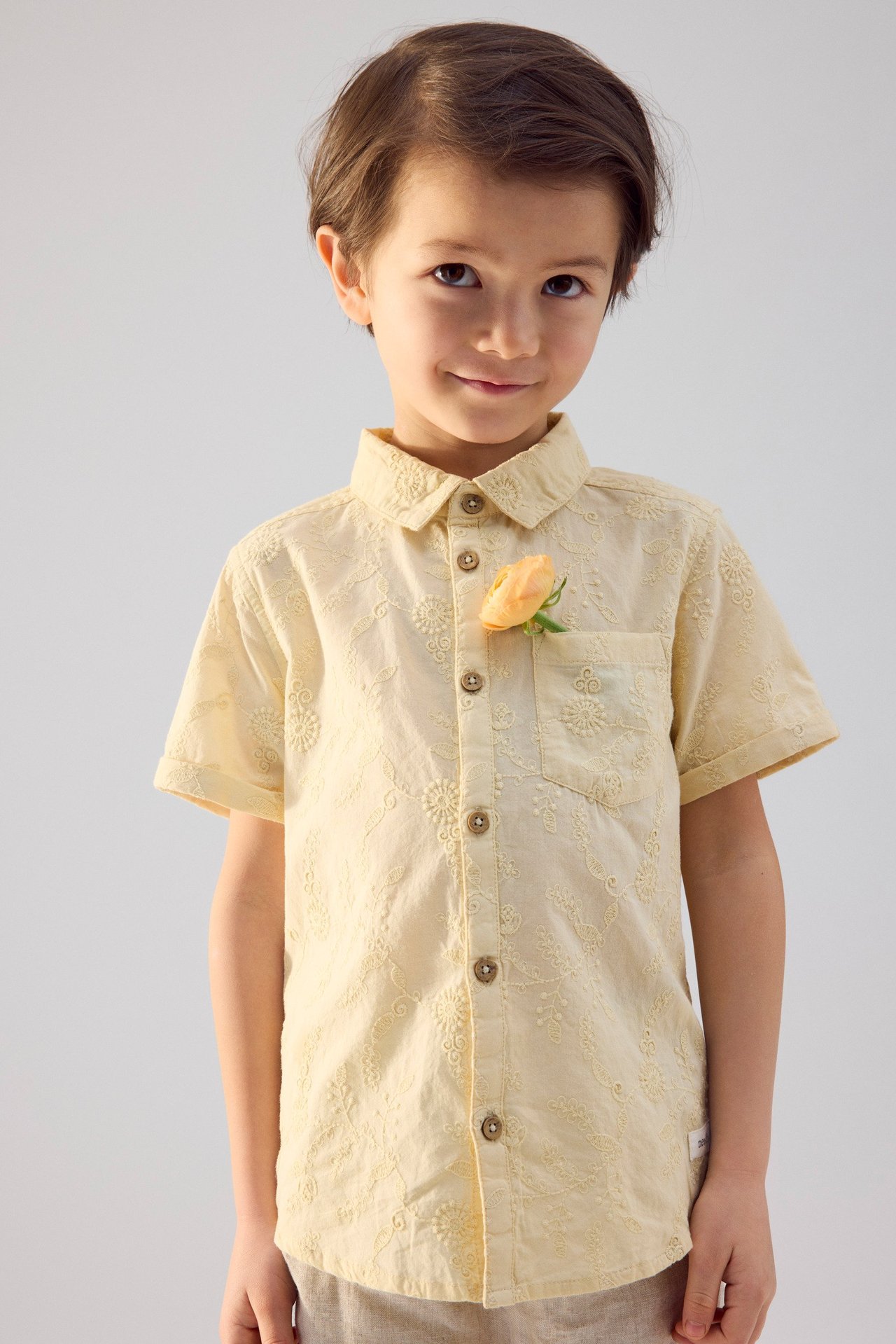 Vorderansicht eines Kindes in einem gelben kurzärmeligen Baumwollhemd mit Blumenstickerei und Blume in der Tasche.