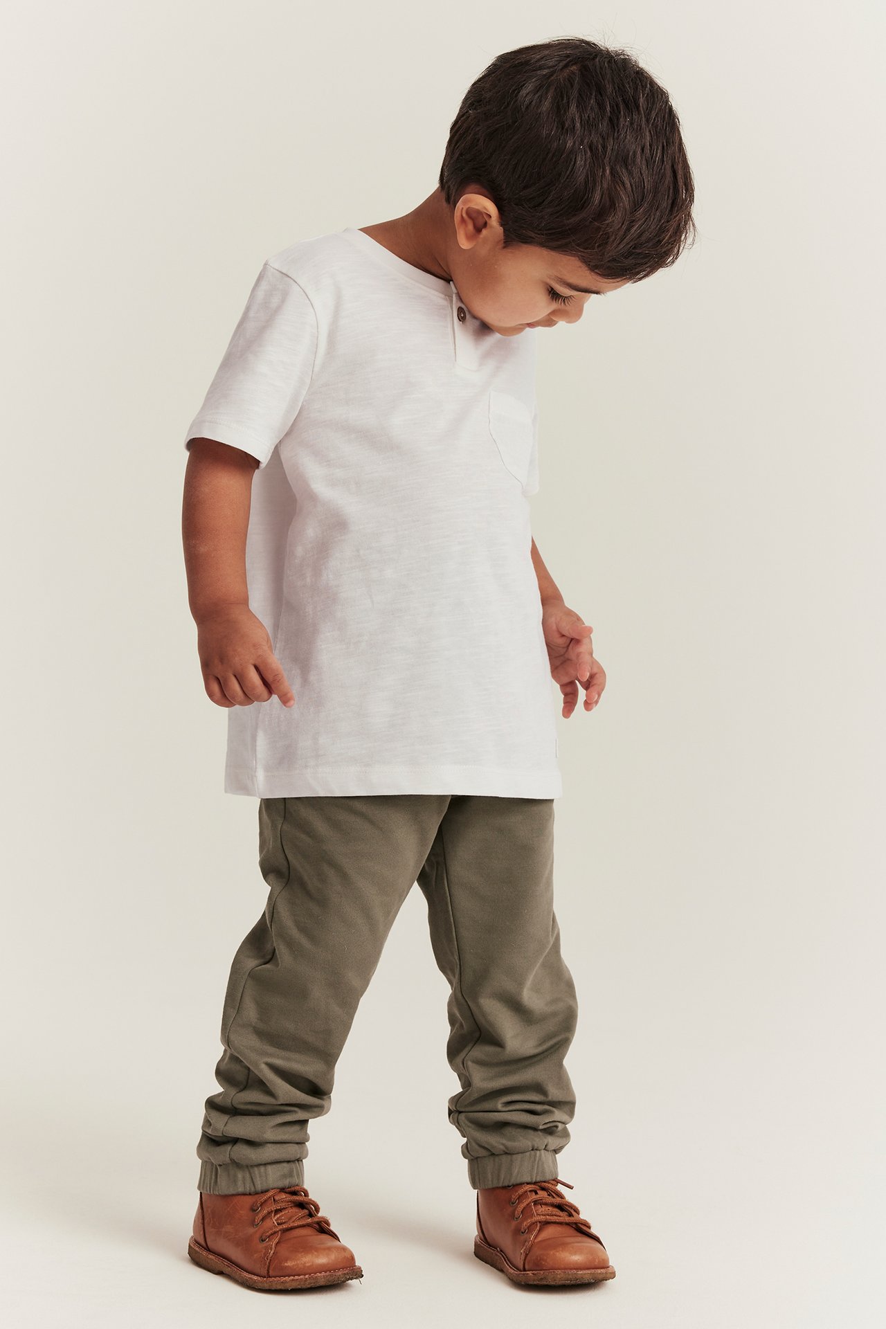 Vorderansicht: Kind in off-weißem Slub-T-Shirt mit Brusttasche, olivgrüner Jogginghose, braunen Stiefeln.
