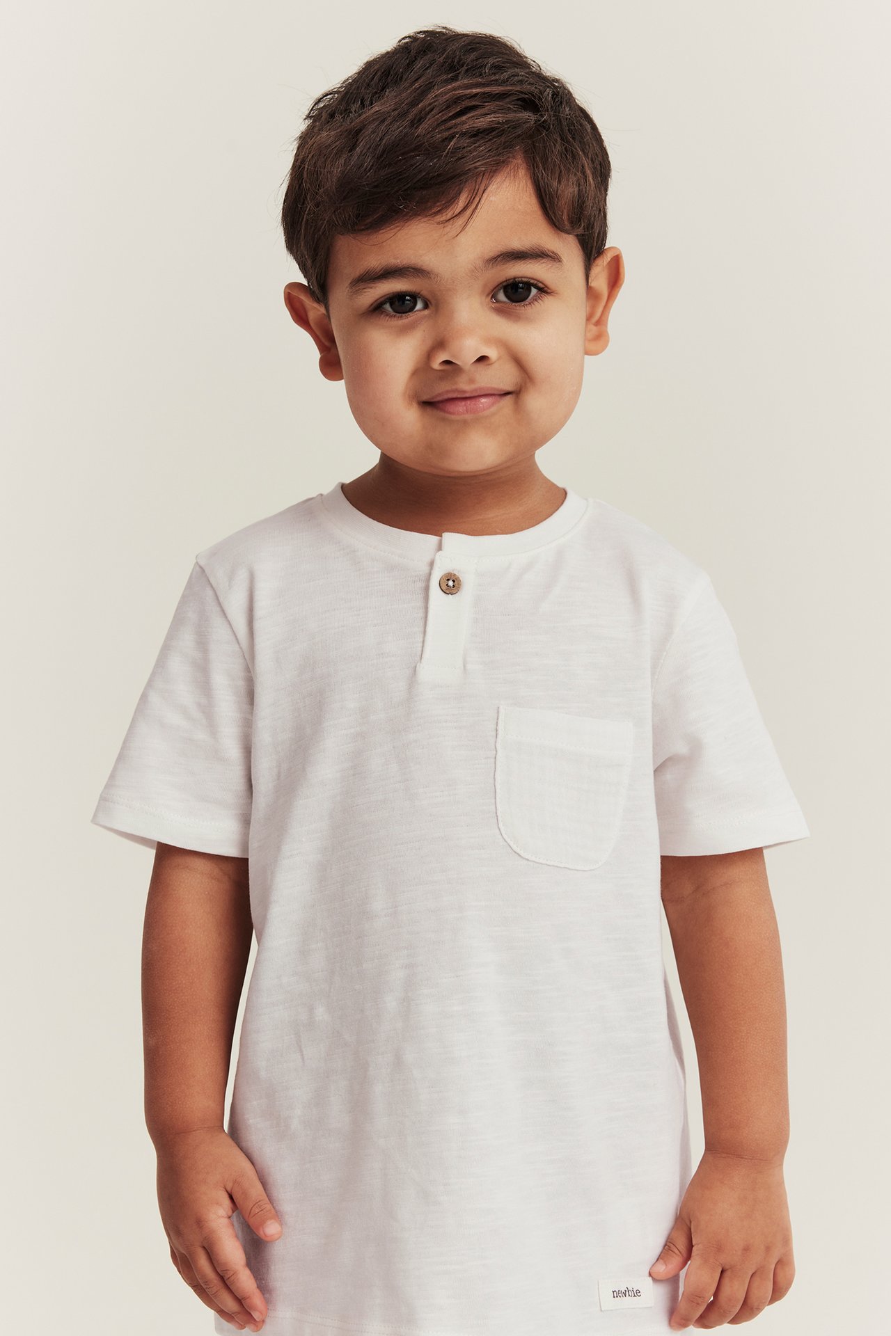 Von vorne gesehen, ein Kind in einem weißen, kurzärmeligen Baumwoll-T-Shirt mit Knopf und Brusttasche.