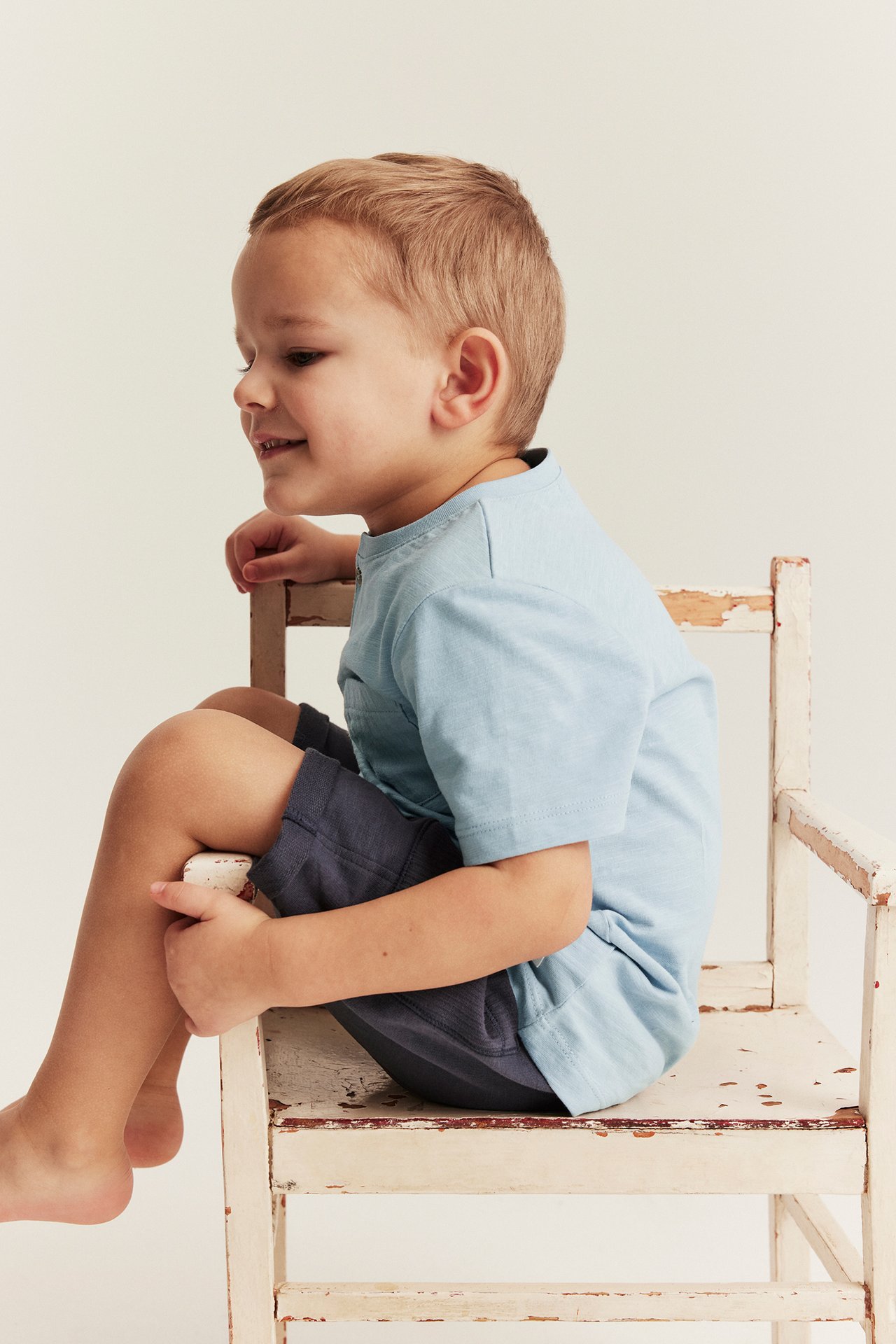 Seitenansicht: Kind in hellblauem Slub-Jersey-T-Shirt mit Brusttasche, Knopf und dunkelblauen Shorts.