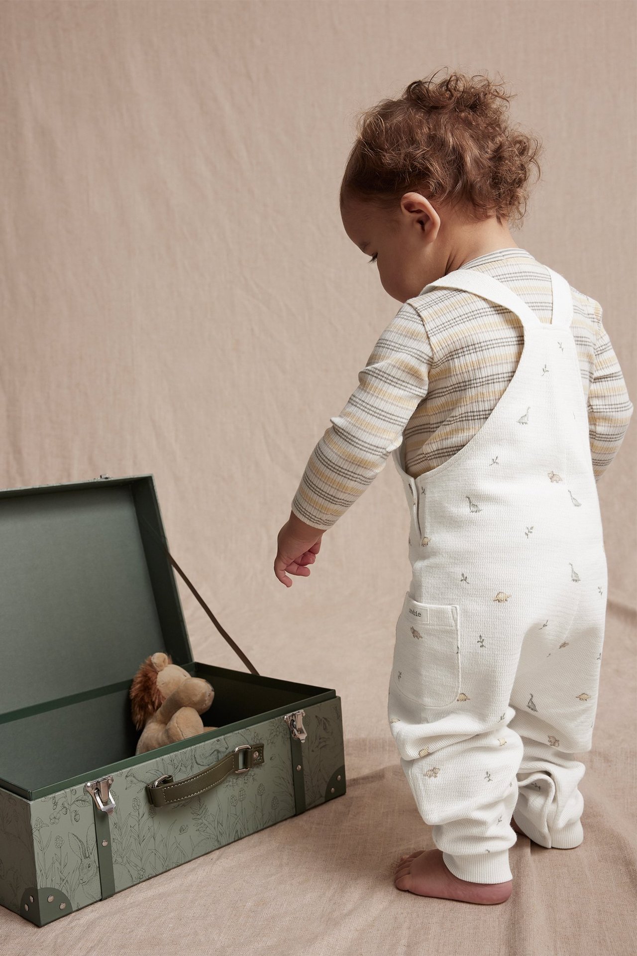 Rückansicht eines Babys in cremeweißer Latzhose mit Dinosaurier-Muster über gestreiftem Langarmshirt, blickt in grünen Koffer.