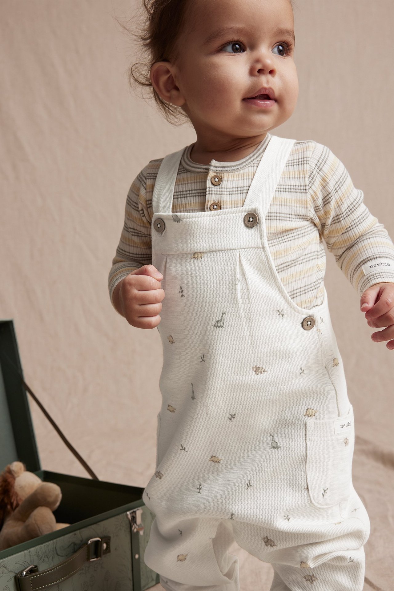 Vorderansicht eines Babys in cremeweißen Latzhosen mit Dinosaurier-Print über einem gestreiften Langarmshirt.
