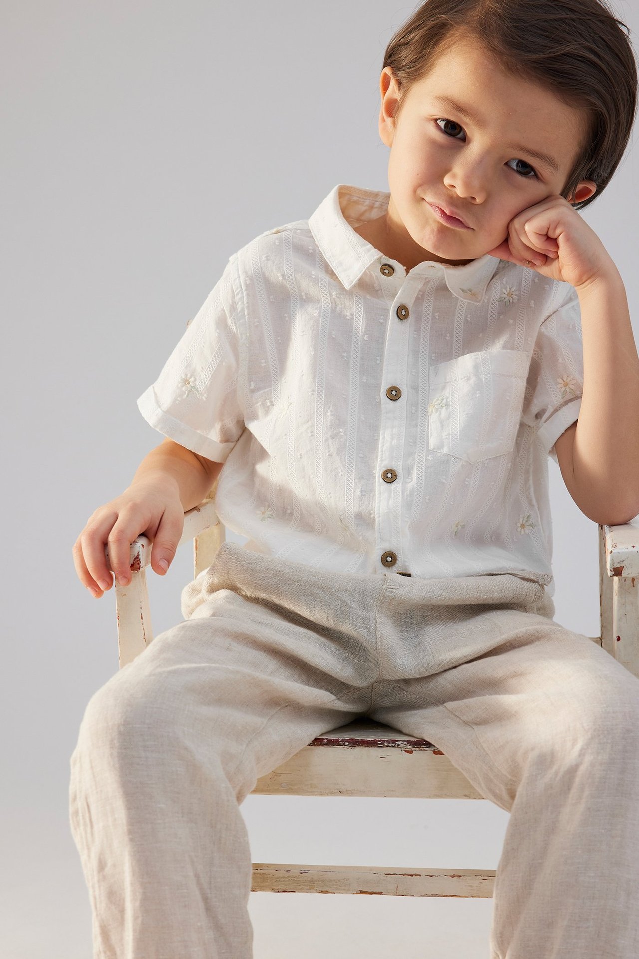 Vorderansicht eines Kindes in einem cremeweißen kurzärmeligen Baumwollhemd mit Blumenstickerei und hellbeiger Hose.