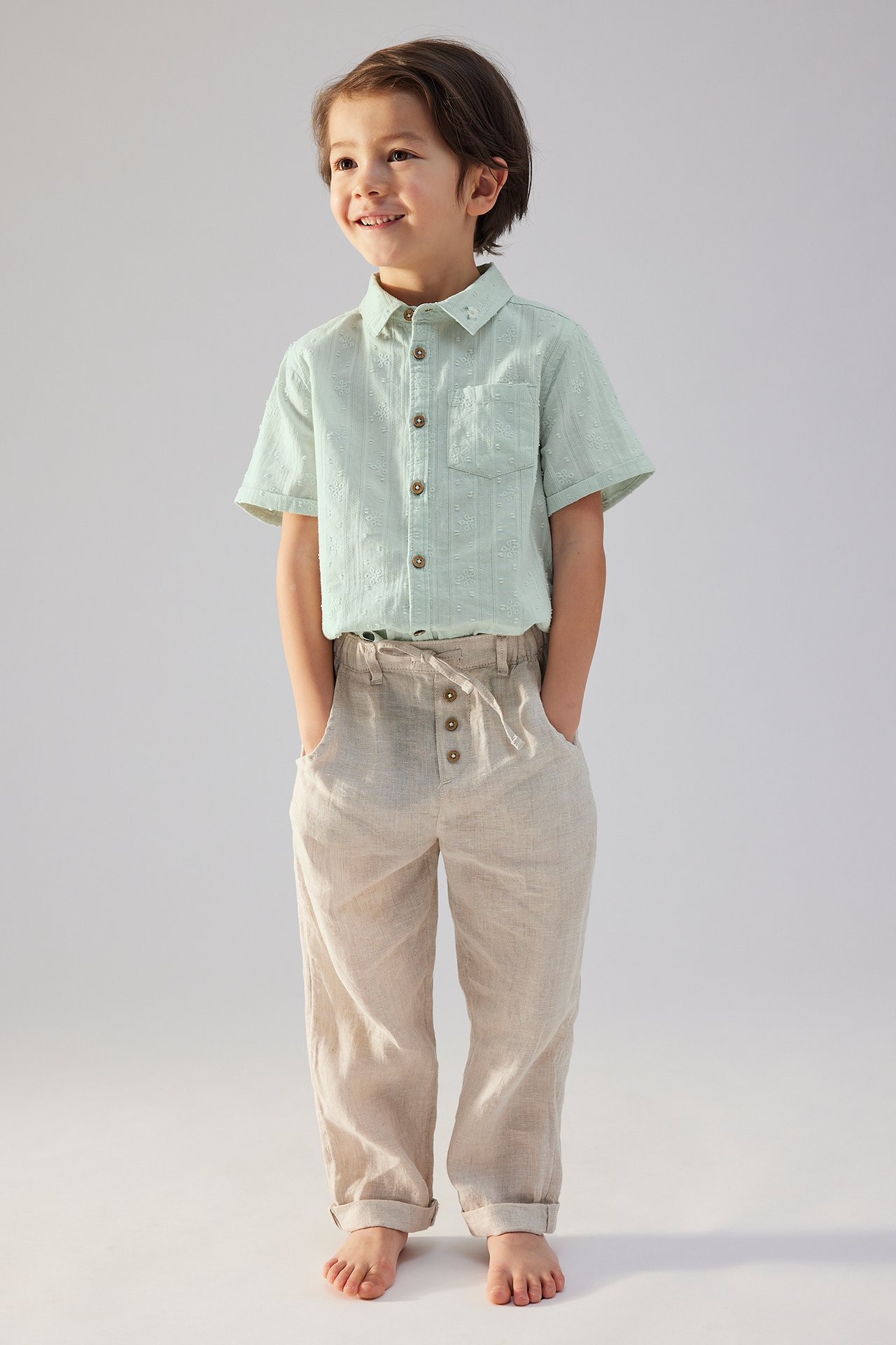 Front view of a kid in a light green embroidered short-sleeved cotton shirt and light beige linen trousers.