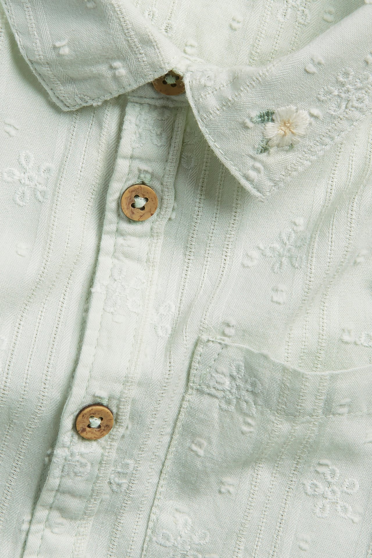 Close-up of a light green cotton short-sleeved shirt with embroidered details, collar, and wooden buttons.
