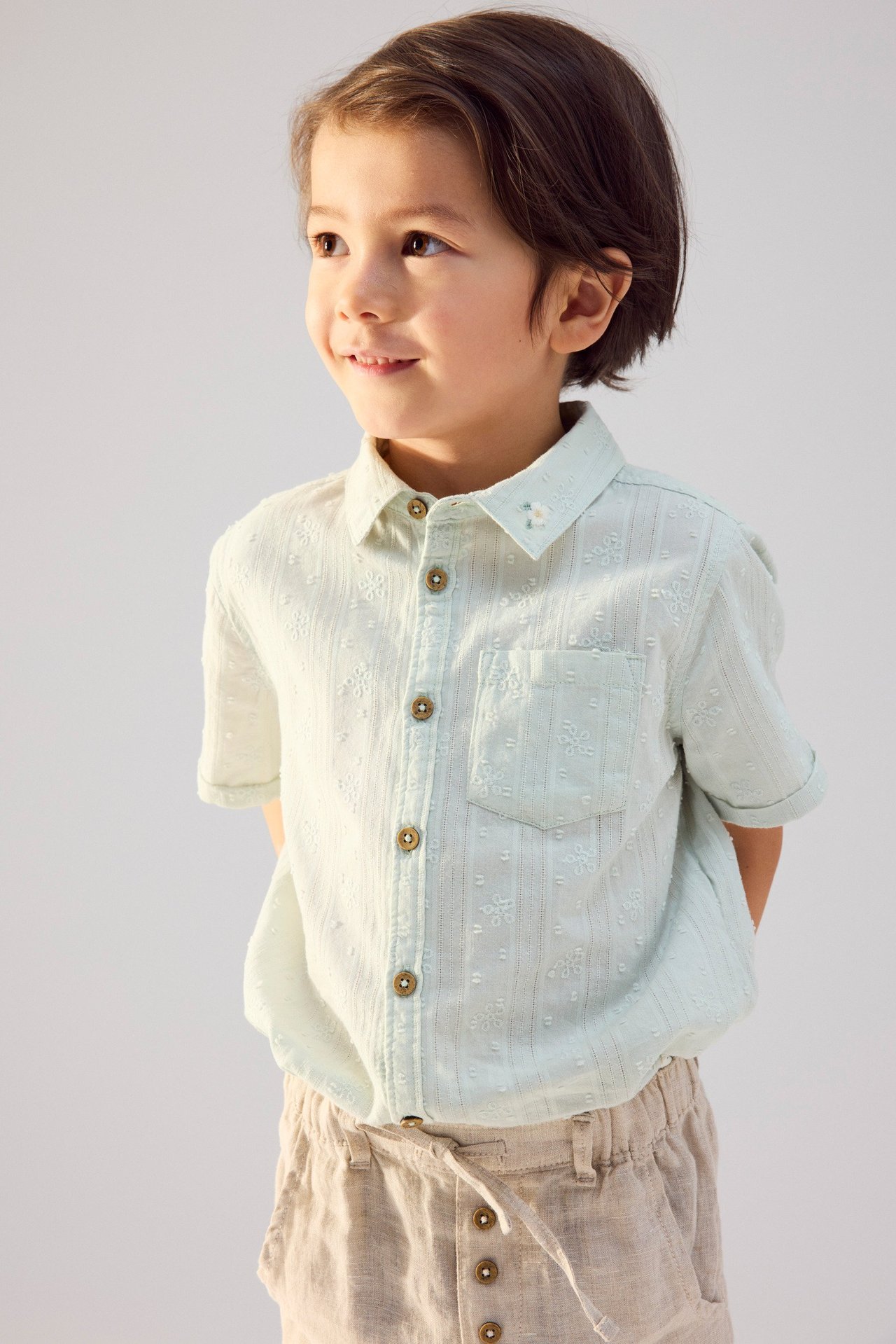 Vorderansicht eines Kindes in hellgrünem kurzärmeligem Baumwollhemd mit Stickerei und beigen Leinen-Shorts.