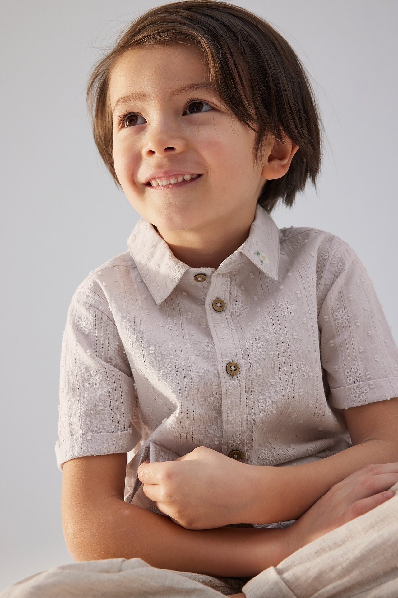 Vorderansicht: lächelndes Kind in helllila kurzärmeligem Baumwollhemd mit weißer Stickerei, Knöpfen und heller Hose.