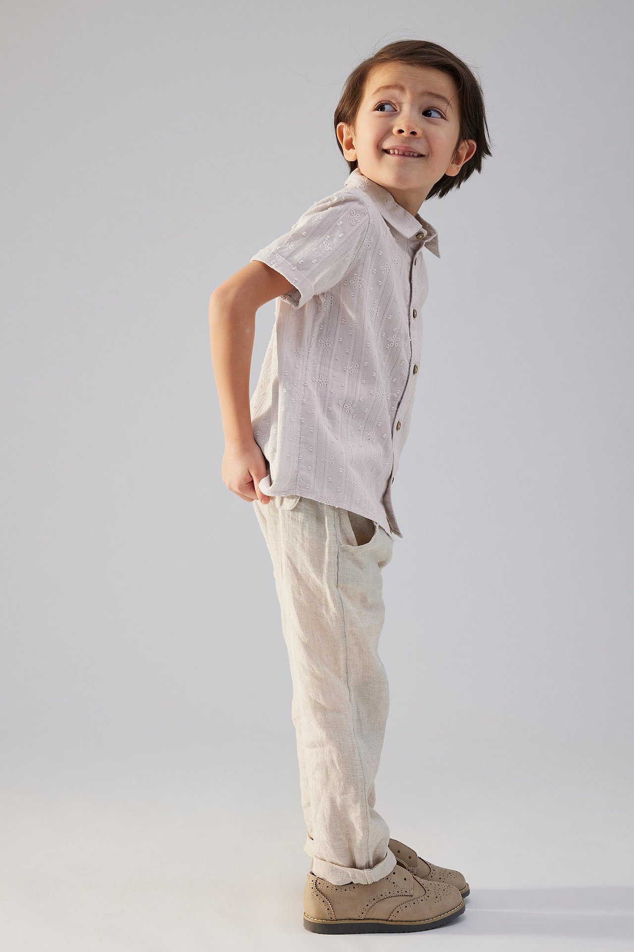 Seitenansicht eines Kindes in einem helllila bestickten Kurzarmhemd, hellbeiger Hose und hellbraunen Schuhen.