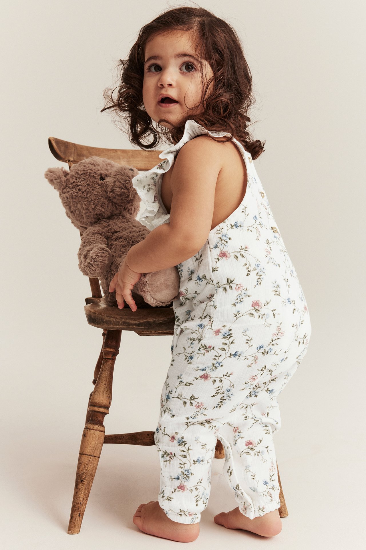 Seitenansicht eines Babys in cremeweißer geblümter Baumwoll-Latzhose mit Rüschenträgern, hält einen braunen Teddy.