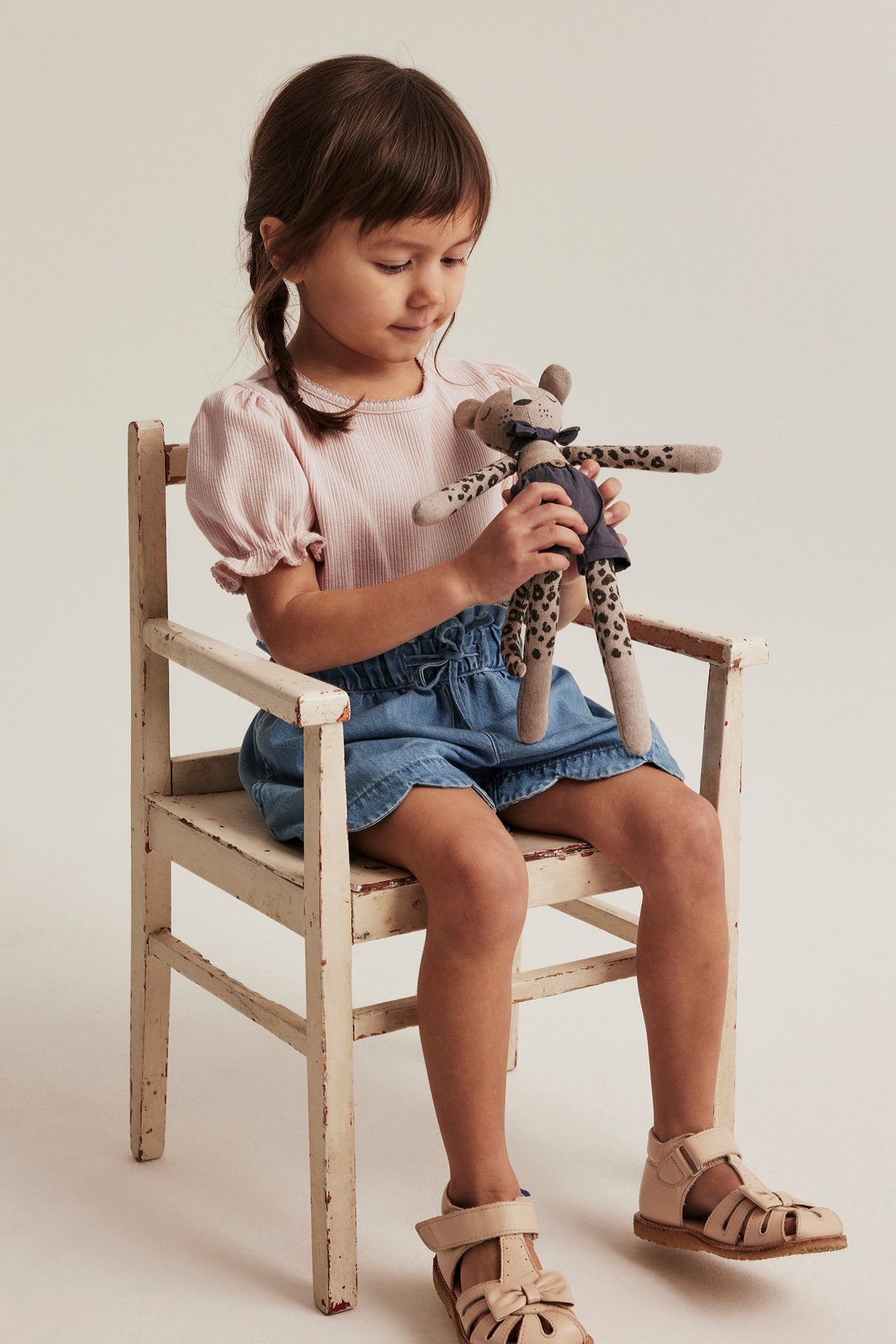 Front view: Kid in light pink ribbed puff-sleeve top, blue shorts, beige sandals, holding a leopard stuffed animal.