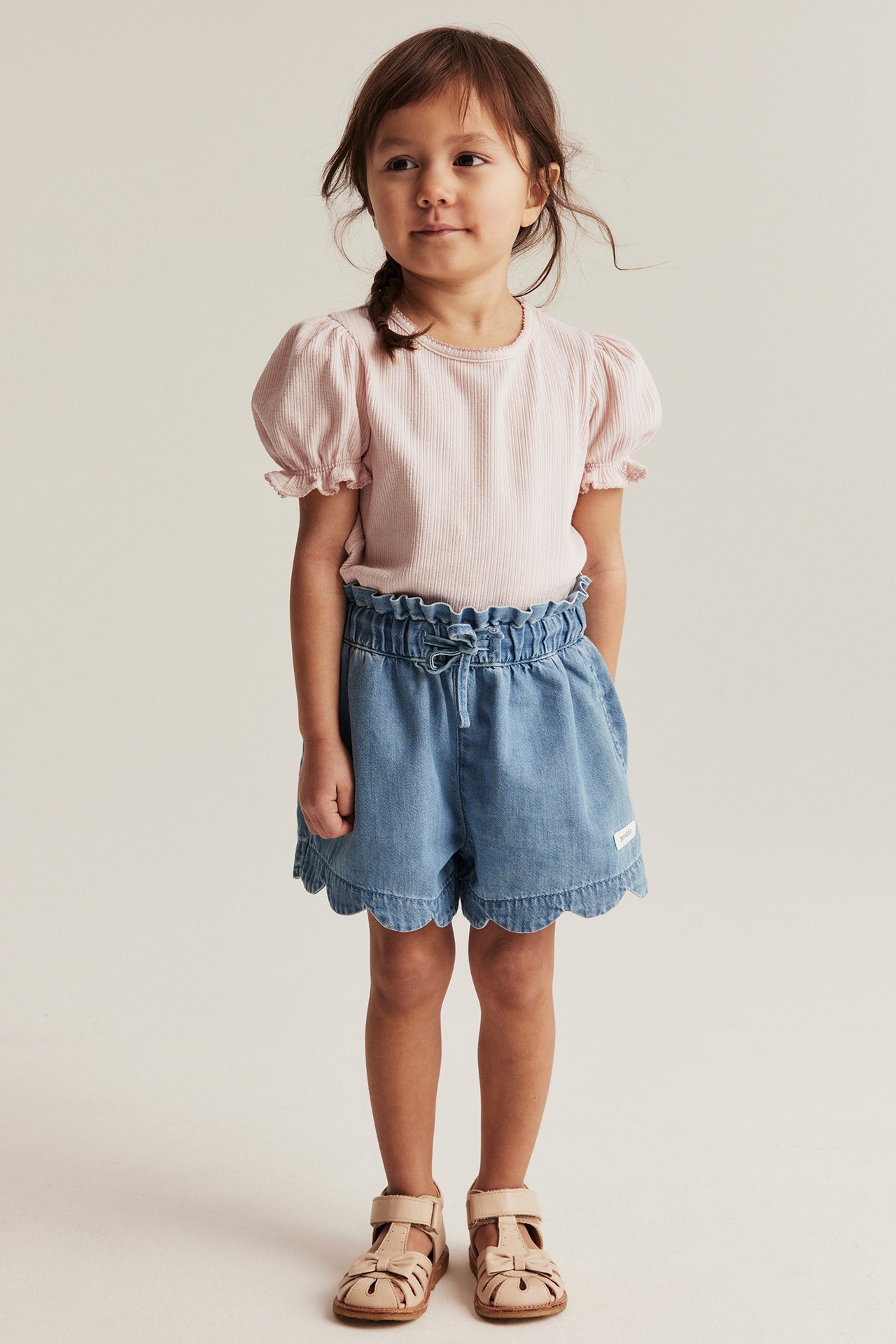 Front view of a kid wearing a light pink ribbed jersey top with puff sleeves, blue denim shorts, and beige sandals.
