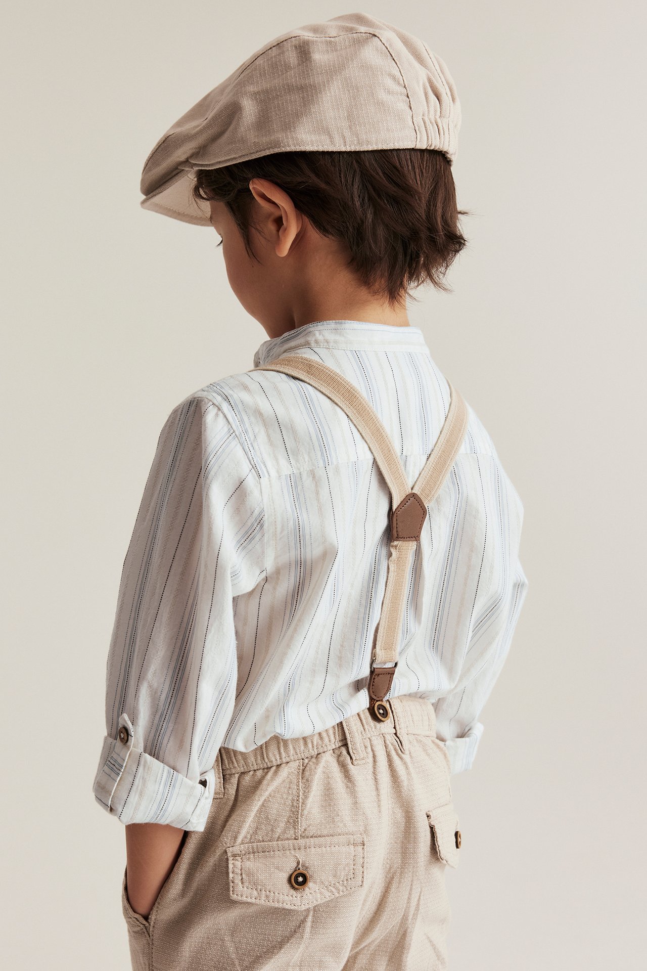 Rückenansicht Kind: gestreiftes off-white Baumwollhemd, beige Hosenträger, beige Hose und Schiebermütze.