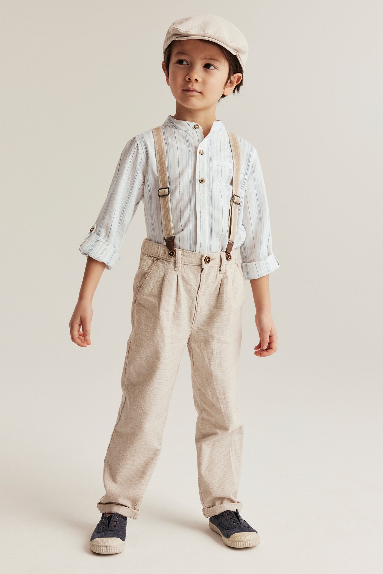 Kind trägt off-weißes gestreiftes Baumwollhemd, beige Faltenhose mit Hosenträgern, beige Mütze und blaue Schuhe.
