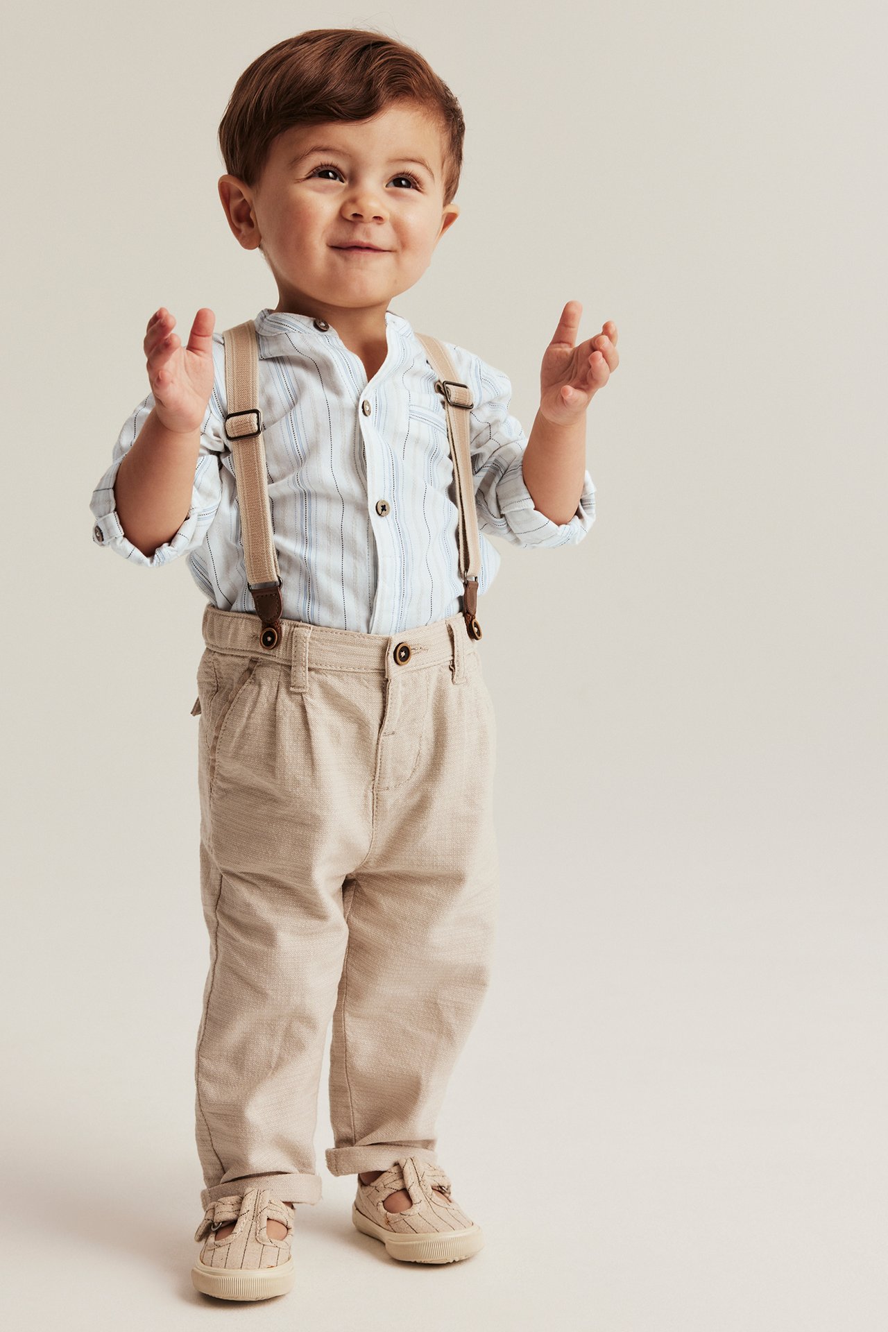 Front view: baby boy in off-white, light blue striped cotton shirt body, beige trousers, suspenders, and shoes.