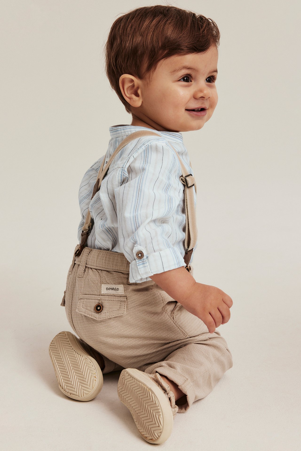 Side view of a baby boy wearing an off-white striped long-sleeved shirt body, beige trousers, suspenders, and sneakers.