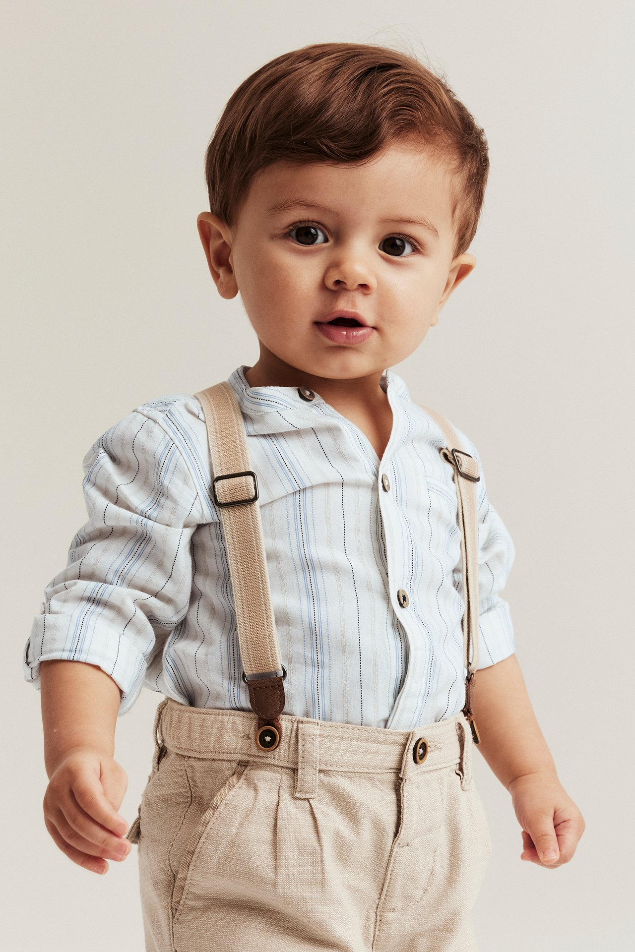 Front view of a baby wearing an off-white striped cotton shirt-body with beige trousers and suspenders.
