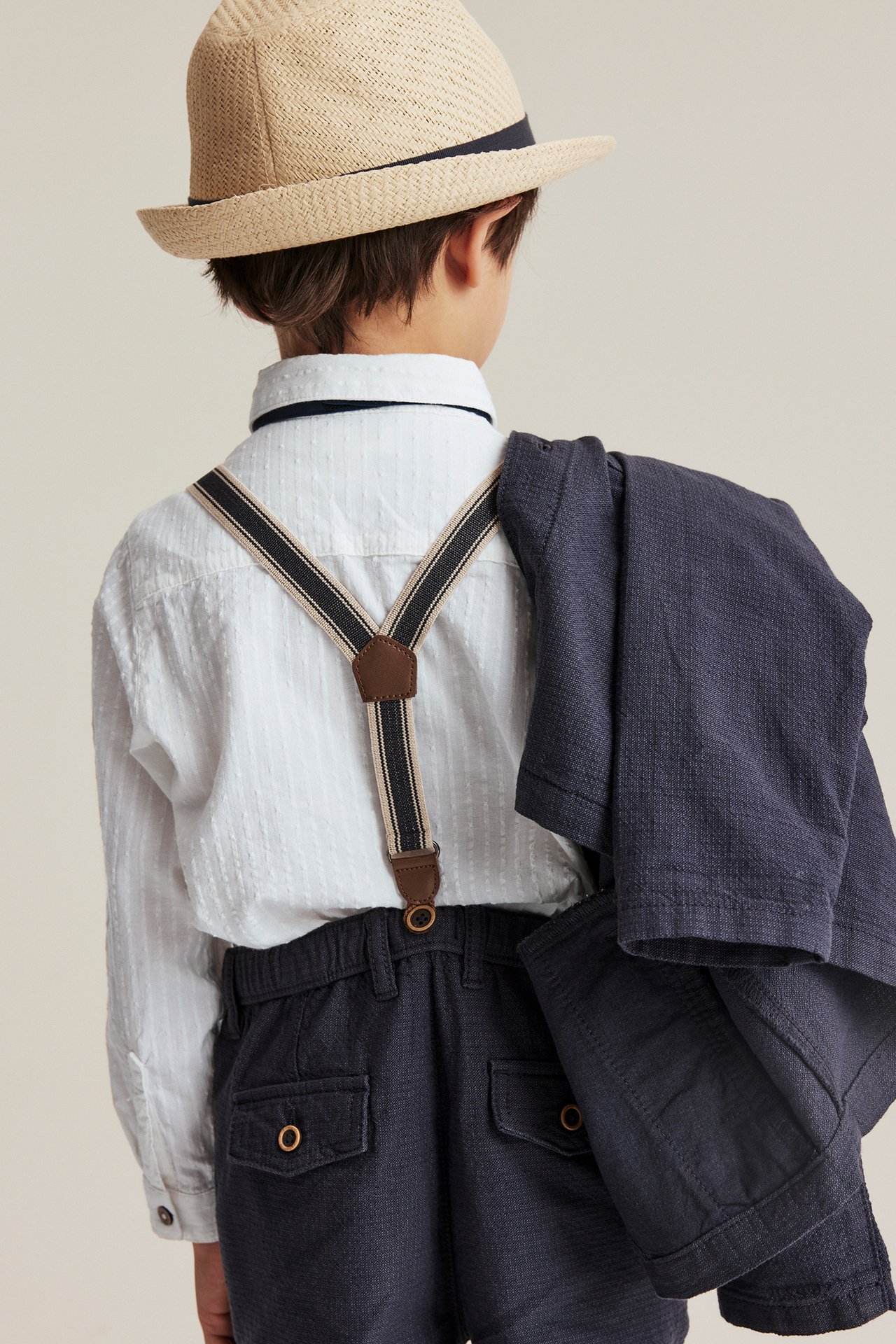 Kid's back view: textured off-white shirt, dark blue shorts, striped suspenders, straw hat, and draped dark jacket.