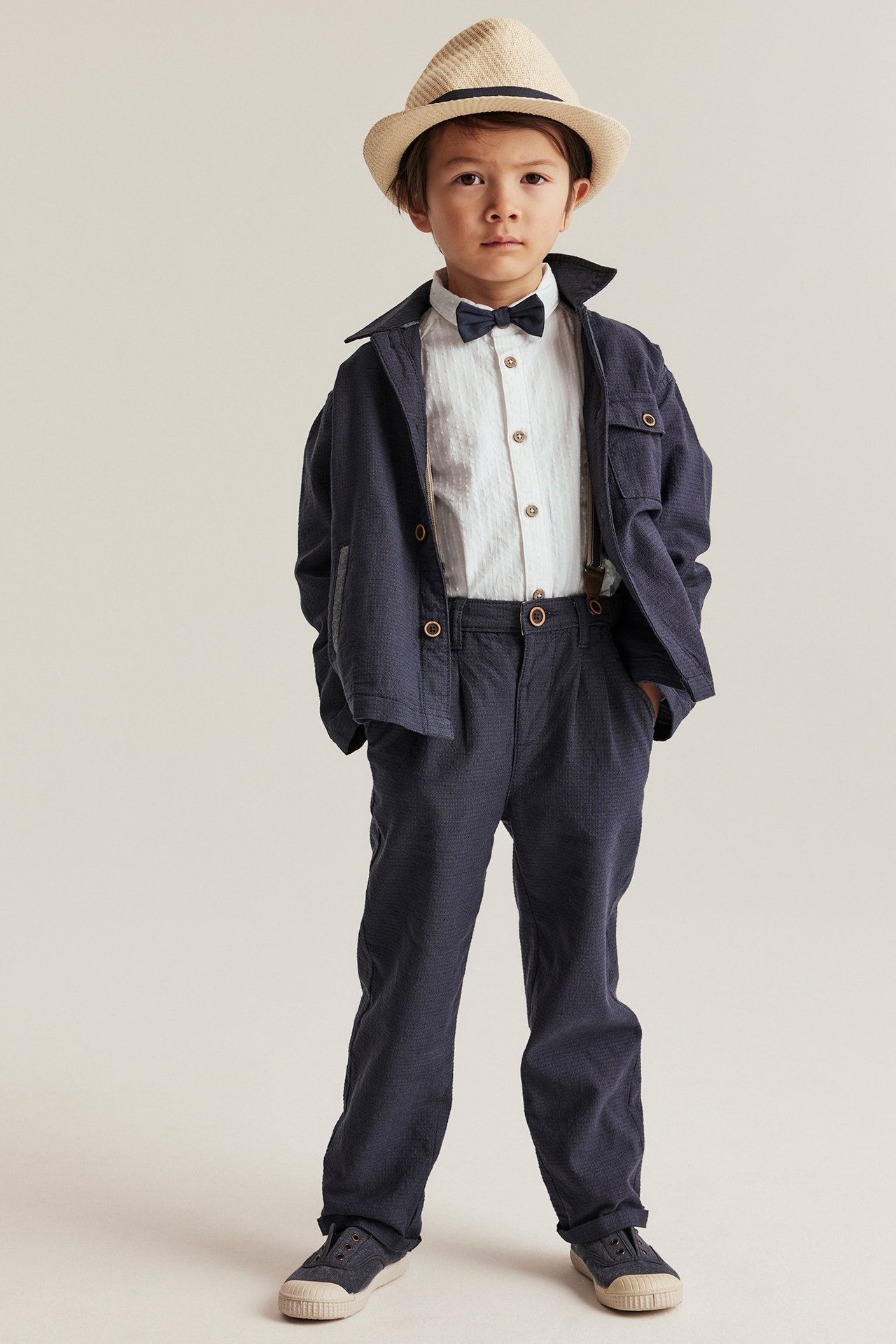 Front view of a kid in an off-white woven shirt, dark blue bow tie, jacket, trousers, and a beige hat.
