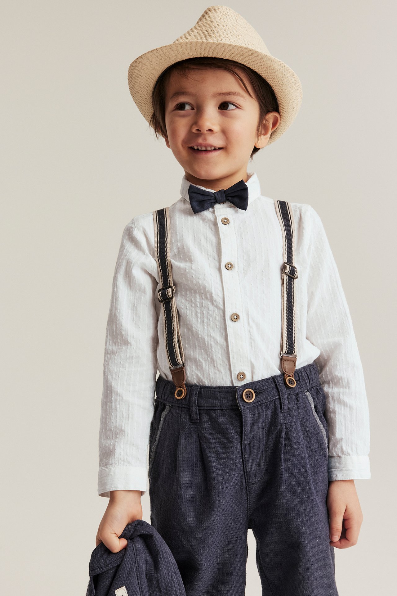 Front view: Kid in offwhite woven shirt with dark blue bow tie, dark blue trousers, suspenders, straw hat, holding cap.