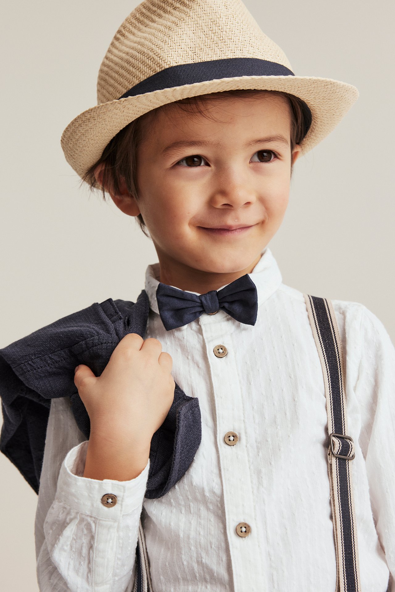 Front view of a kid in an off-white woven shirt, dark blue bow tie, striped suspenders, and straw hat.