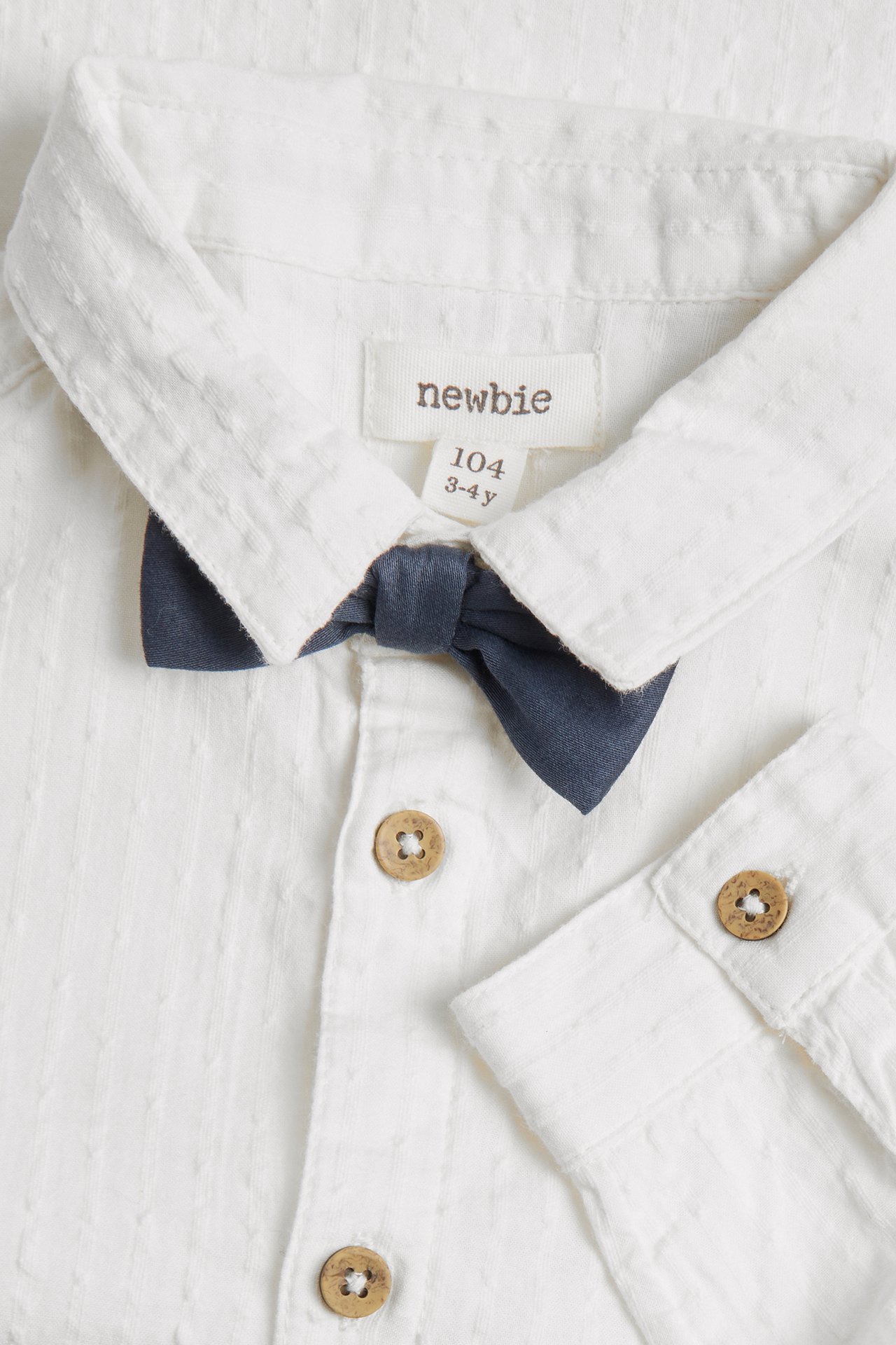 Close-up of a kid's off-white cotton shirt with a dark blue detachable bow tie and wooden buttons.
