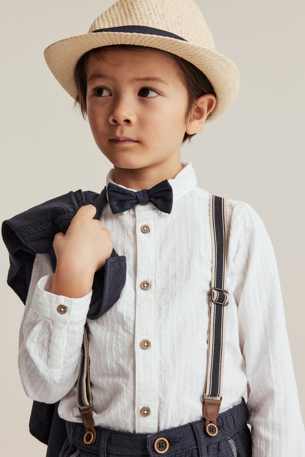 Front view of a kid, offwhite woven shirt, dark blue bow tie, striped suspenders, and straw hat.