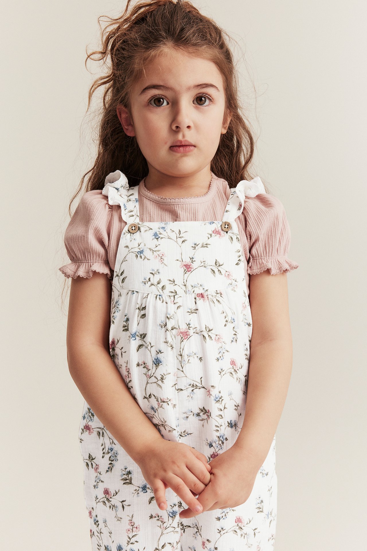 Vorderansicht eines Kindes in cremeweißen geblümten Latzhosen mit Rüschen über einem rosa gerippten Kurzarmshirt.