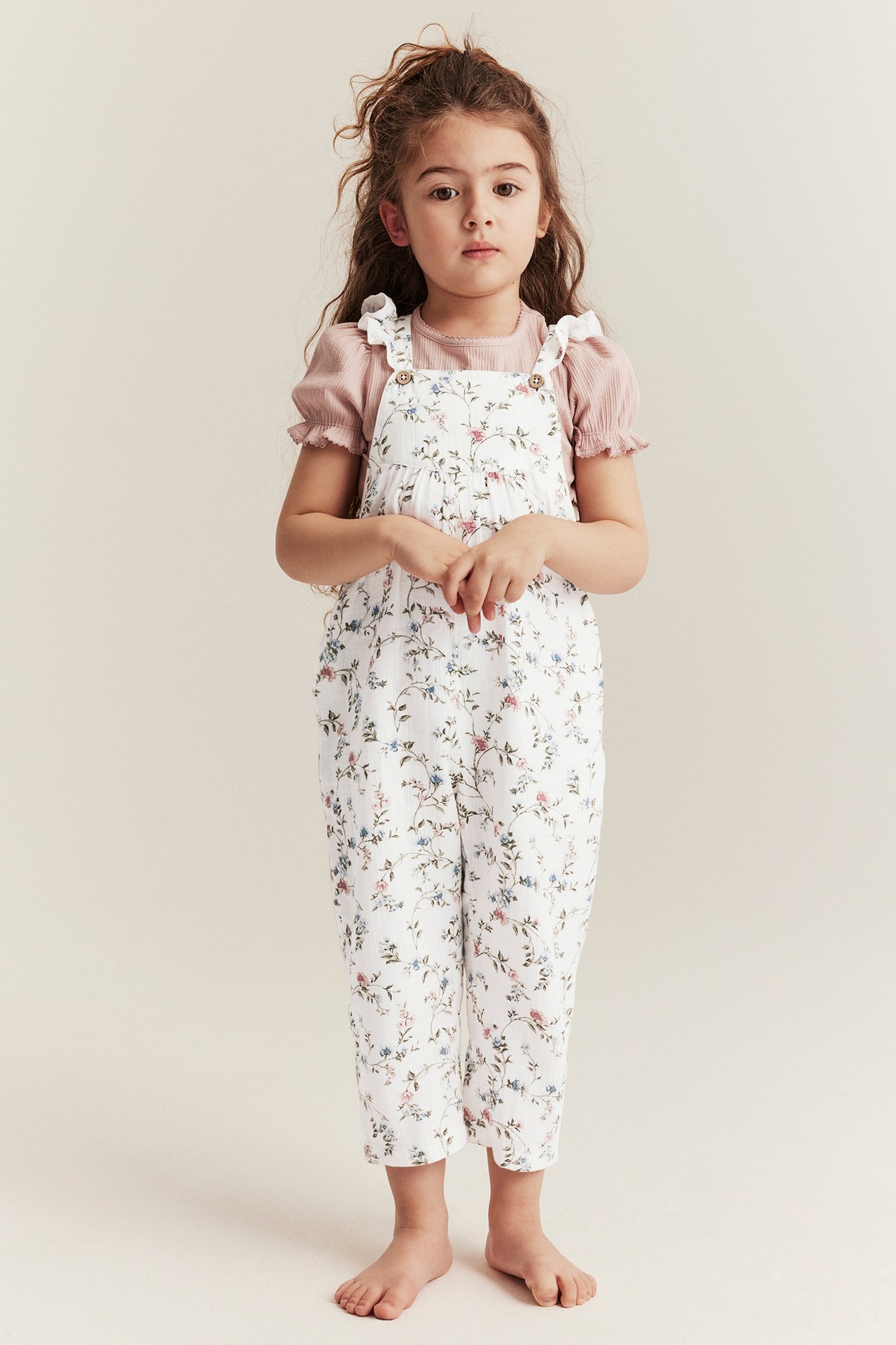 Vorderansicht eines Kindes in offwhite geblümter Latzhose mit Rüschenträgern über einem rosa kurzärmeligen Oberteil.