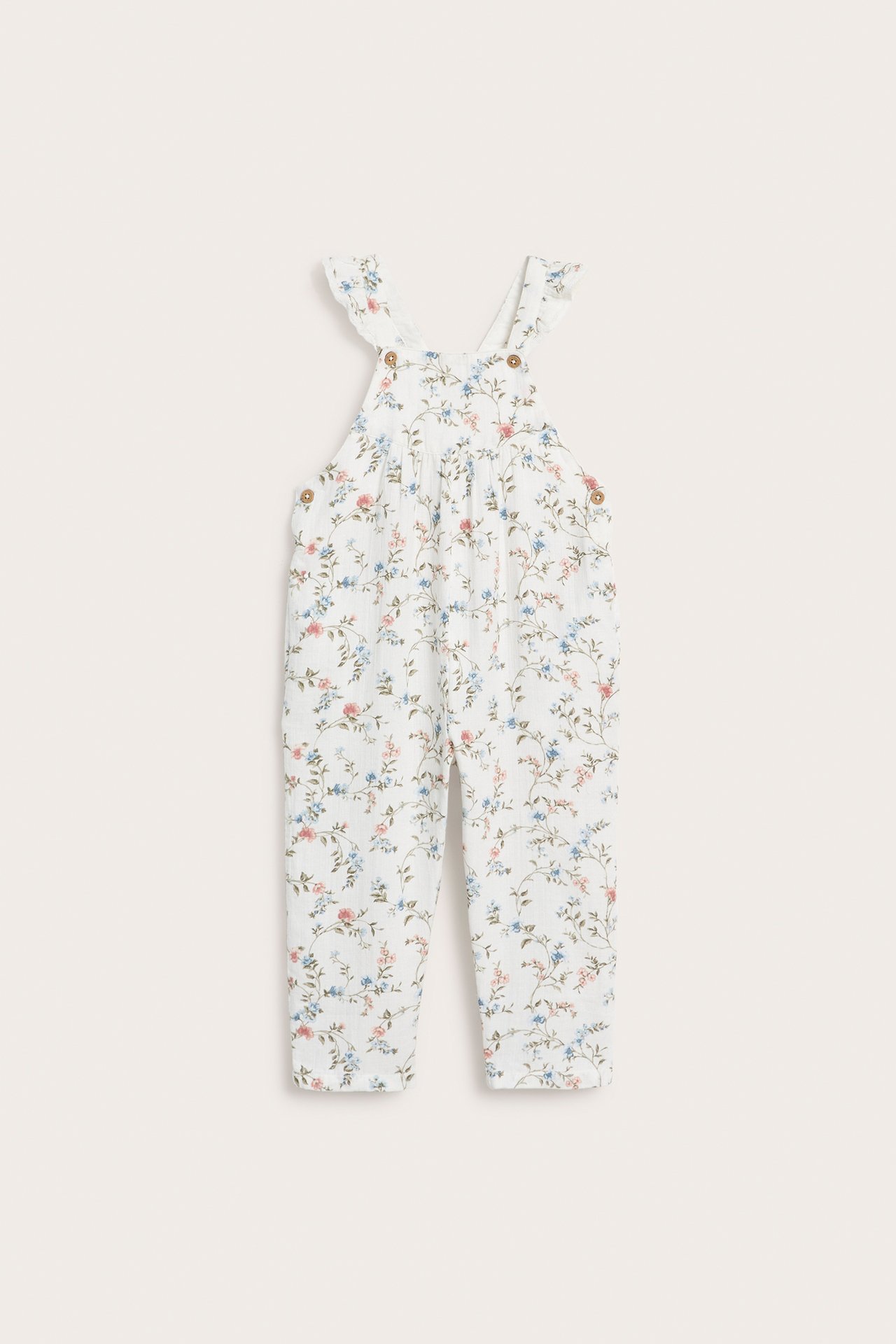 Vorderansicht: Kinder-Latzhose aus cremefarbener Baumwolle mit Blumenmuster, Rüschen an Trägern, Holzknöpfen.