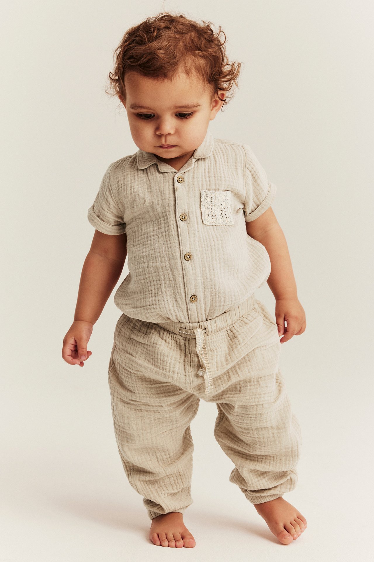 Vorderansicht eines Babys in beiger Musselin-Hose mit Kordelzug und passendem Knopfhemd.