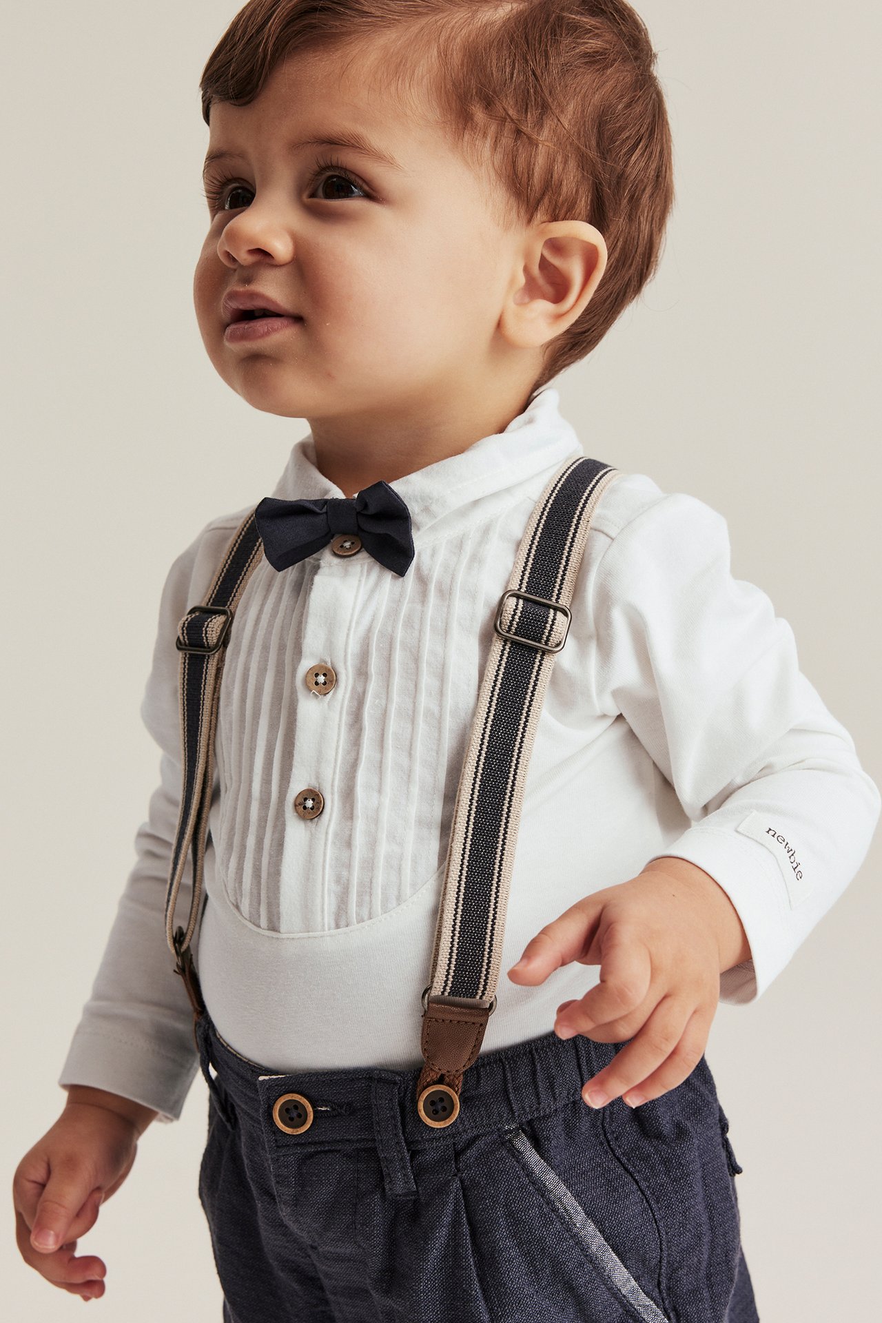 Vorderansicht eines Babyjungen in einem cremefarbenen Langarm-Body, dunkelblauer Fliege, dunkler Hose und Hosenträgern.