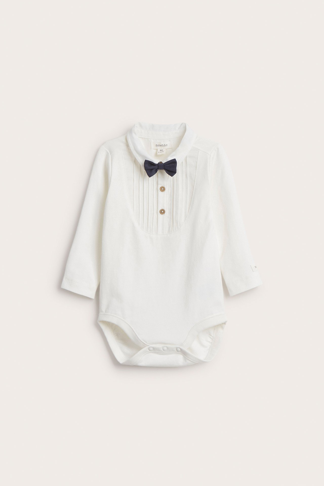 Vorderansicht eines cremeweißen langärmeligen Baby-Bodys mit Kragen, Faltenfront, Knöpfen und dunkelblauer Fliege.