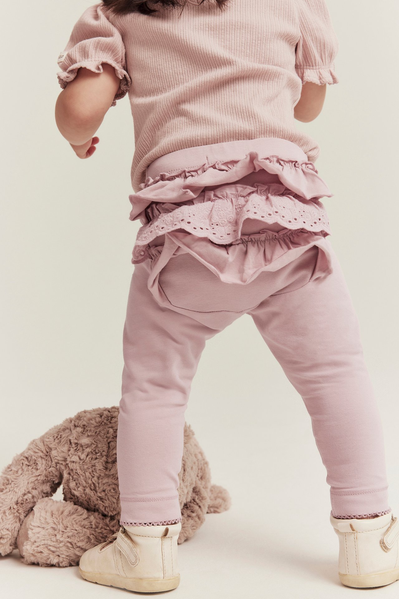 Back view of a baby in pink ruffled lace leggings, a pink ribbed top, and light beige shoes.
